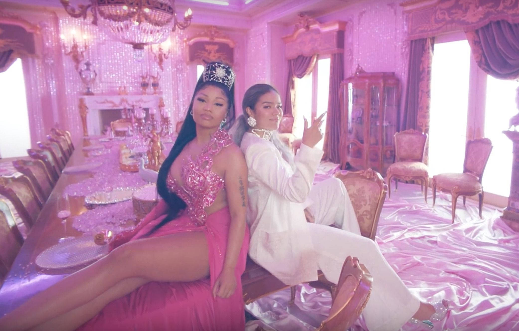 Karol G 與 Nicki Minaj 木桌合照 壁紙 — 兩位女士在一個裝飾華麗的粉紅色豪宅房間裡，閃爍著水晶吊燈和豪華家具，迷人地擺姿勢。
