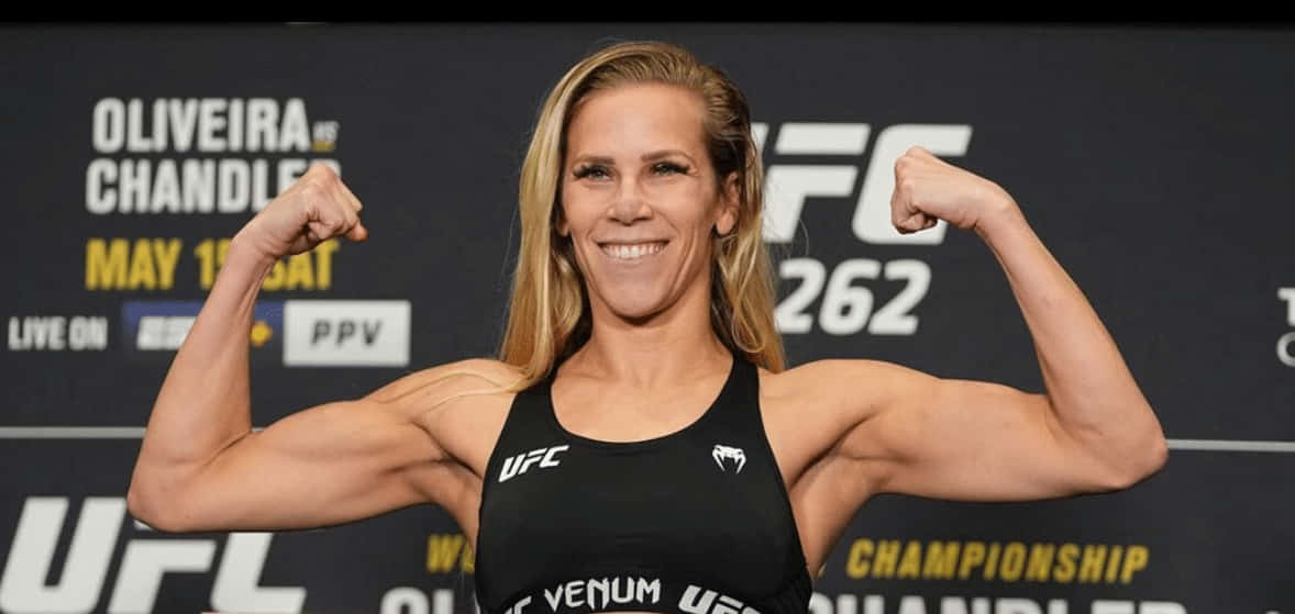 Katlyn Chookagian 開心展示肌肉 壁紙 — 一位微笑的女性UFC拳手在稱重活動中彎曲她肌肉發達的手臂，身後有冠軍品牌標誌。