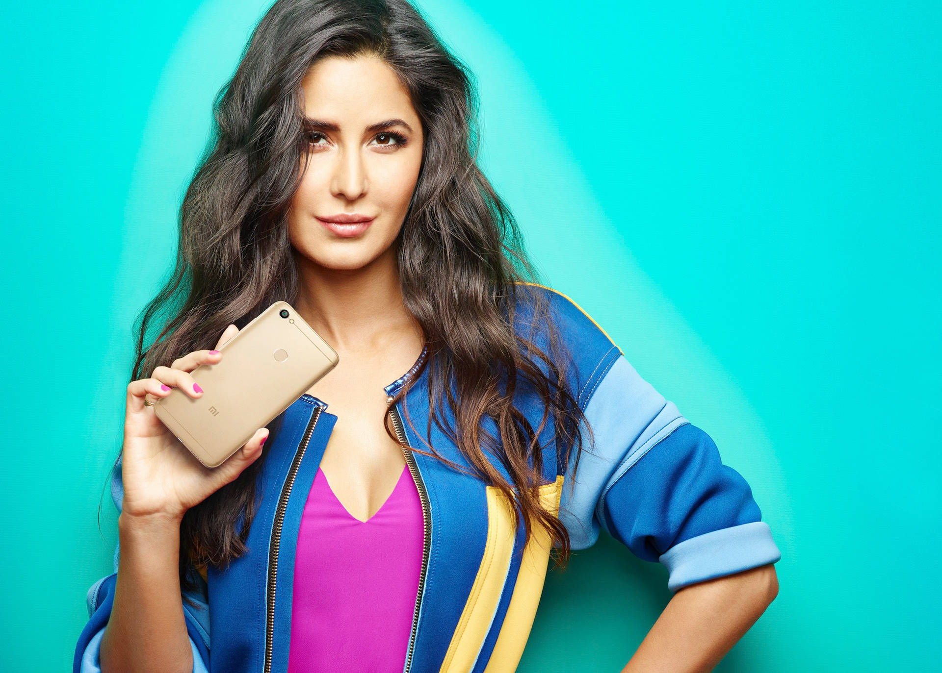 Katrina Kaif 智能手機品牌拍攝 壁紙 — 一位長黑髮的女性在充滿活力的青綠色漸變背景前拿著金色智能手機。