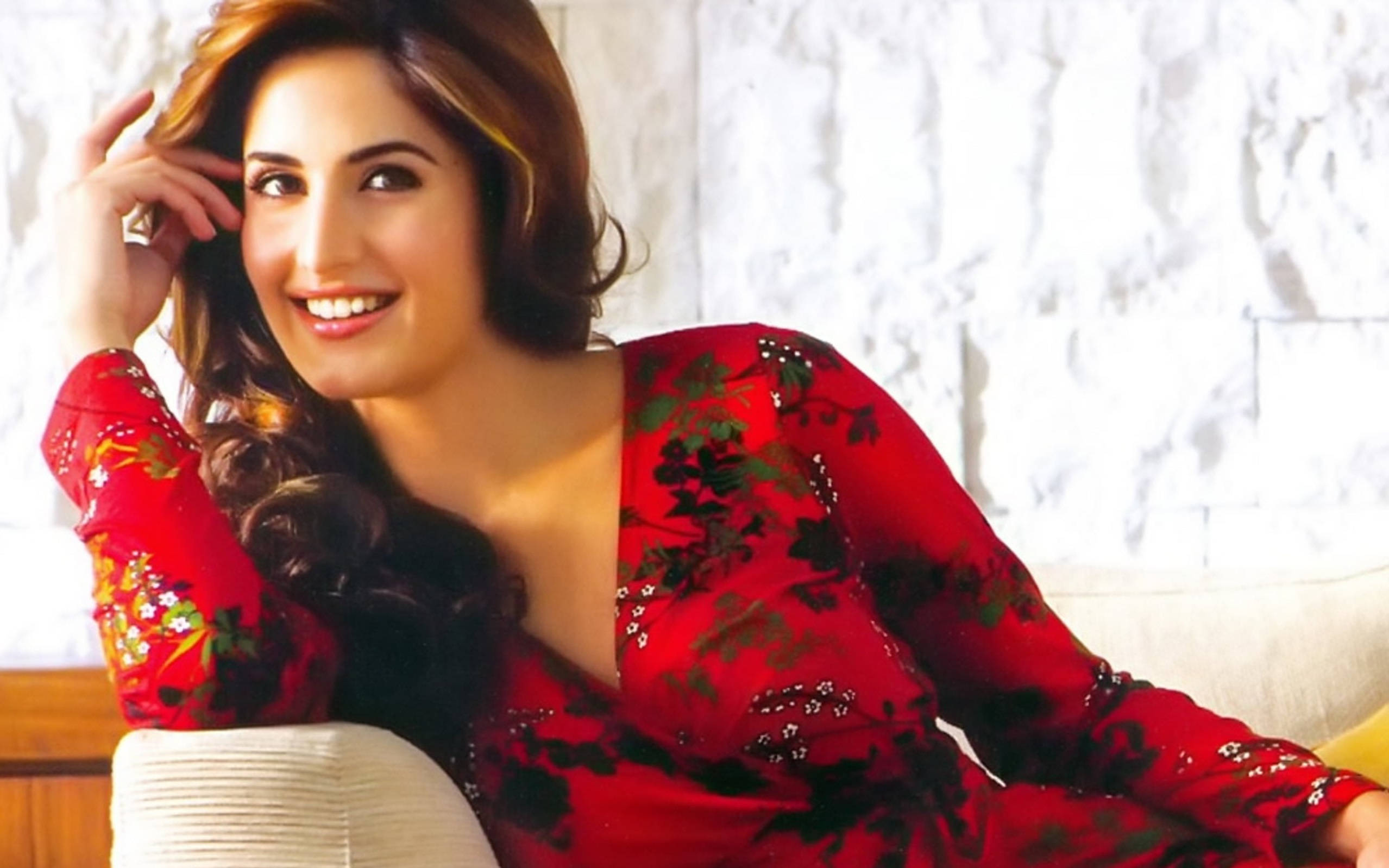 Katrina Kaif 紅色花卉長袖禮服 壁紙 — 一位身穿紅色花卉連衣裙的容光煥發的女性，在白色紋理牆壁前微笑著觸摸她的頭髮。