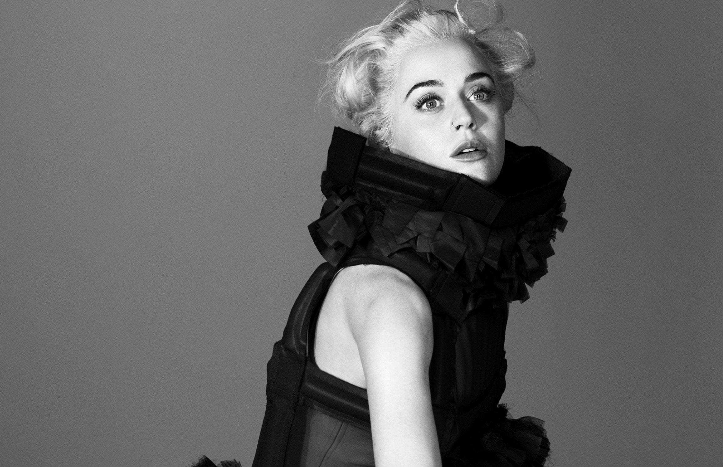 Katy Perry 身穿 Comme Des Garçons 壁紙 — 一個引人注目的黑白肖像，一位金髮蓬鬆的女性，穿著戲劇性的黑色荷葉邊領，背景是純灰色。