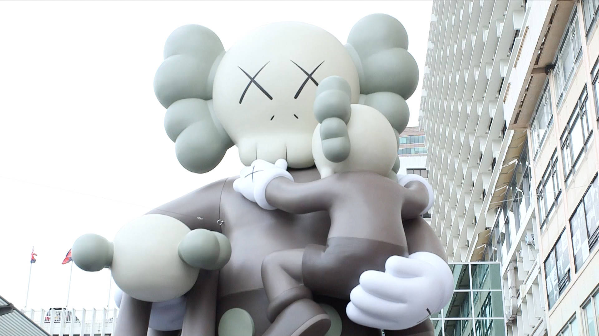 KAWS Clean Slate雕塑 壁紙 — 兩個巨型KAWS公仔喺陰天背景下，喺現代城市建築物前溫柔擁抱。