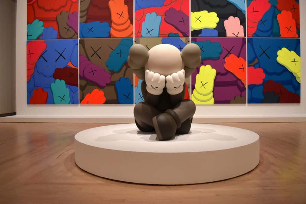 KAWS Companion 粗獷棕熊 壁紙 — AKAWSleepy 雕塑坐在白色底座上，前面是畫廊中色彩繽紛的 KAWS 藝術品網格。