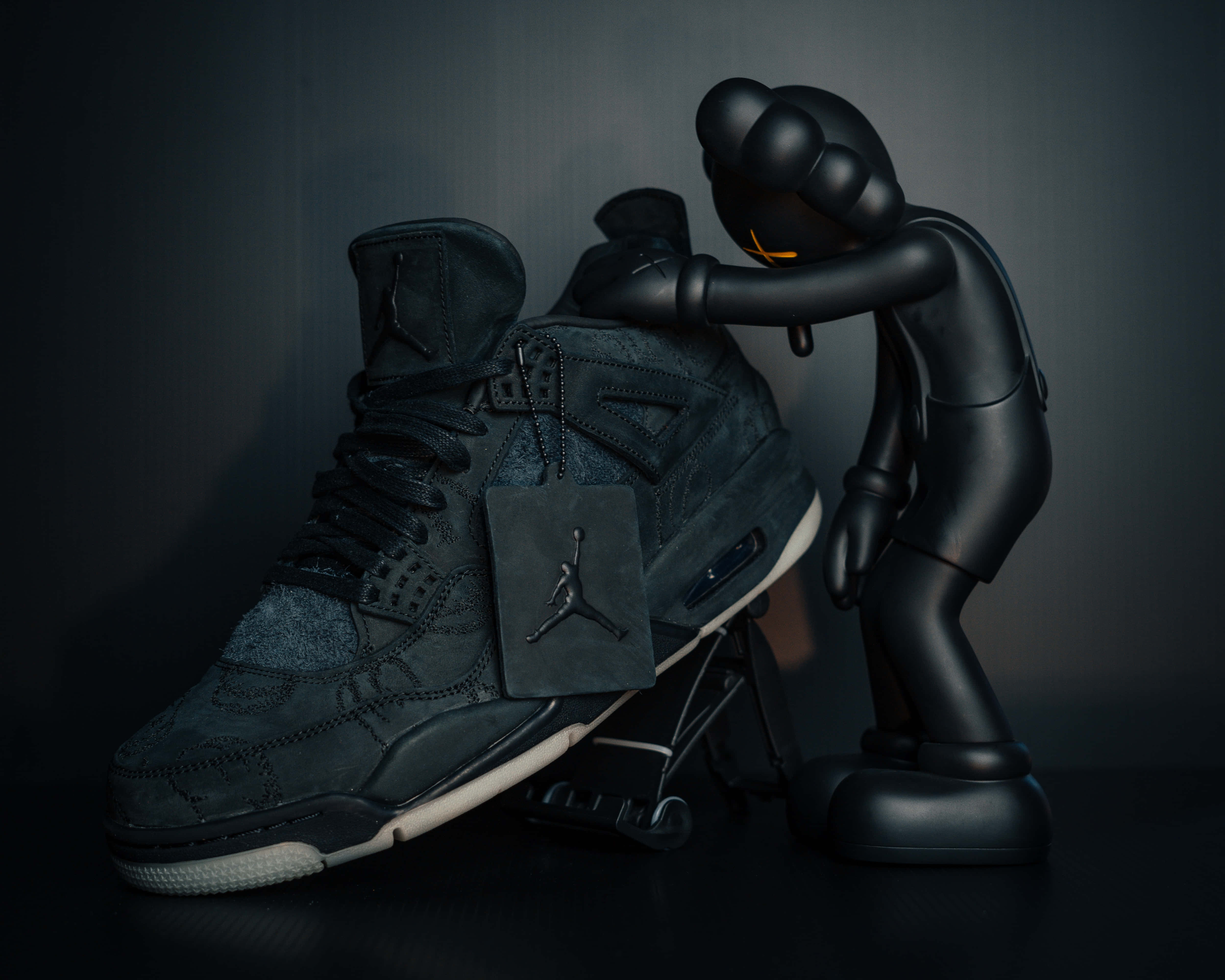 KAWS Companion：經典角色的重新詮釋 壁紙 — 一個亮面黑色KAWS公仔喺深色漸變背景前溫柔咁擁抱住一對黑色Jordan波鞋。