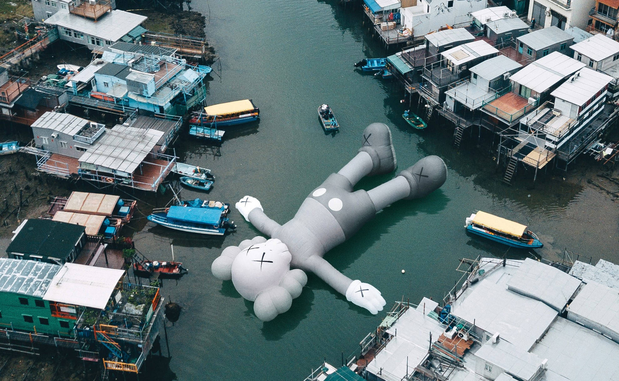 KAWS Companion浮於水面 壁紙 — 一個巨大的灰色KAWS充氣公仔在運河中平靜地漂浮，周圍是高腳屋和小船。
