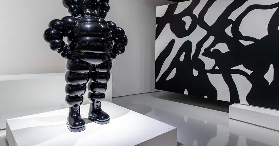 KAWS X Bearbrick 限量版雕塑 壁紙 — 一個光澤的黑色氣泡形雕塑放在白色底座上，位於極簡主義畫廊中，旁邊是一幅大膽的黑白抽象牆面作品。