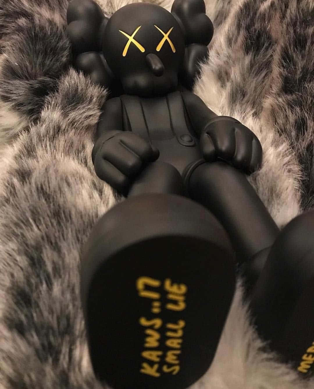 KAWS:X藝術品籠罩城市天際線 壁紙 — 一個黑色KAWS Companion公仔，帶有黃色X眼睛，放在柔軟的灰白色毛絨表面上。