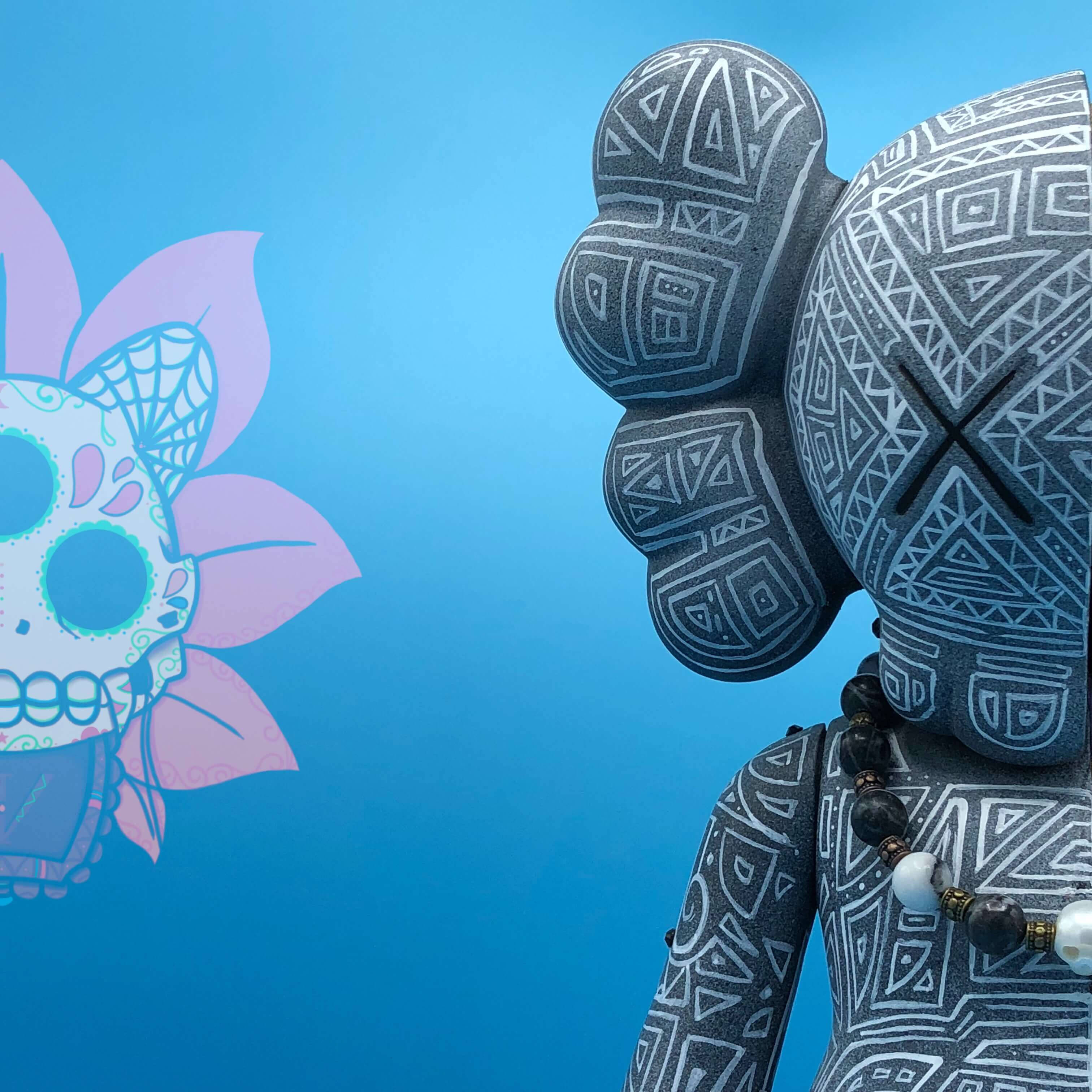KAWS 古代戰士的犧牲 壁紙 — 一個灰色圖案KAWS公仔，帶有黑色X眼睛，在純藍色背景上凝視著一個色彩繽紛的糖骷髏花旁邊。