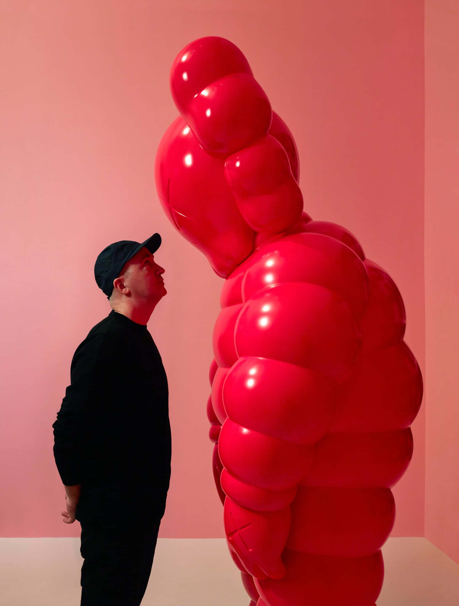 KAWS 在 Louis Vuitton 基金會展覽現場 壁紙 — 一名身穿黑衣的男子敬畏地站在一座高聳的亮紅色氣球雕塑旁，背景是柔和的粉紅色牆壁。
