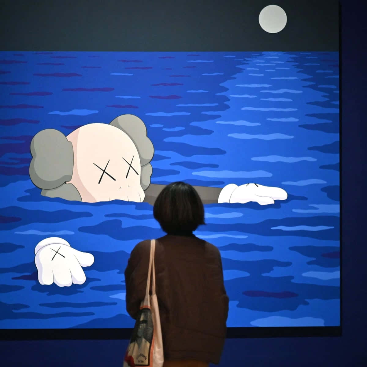 KAWS 在紐約慶祝時光 壁紙 — 一個人正在觀看一個大屏幕，顯示一個卡通風格的KAWS角色在滿月下浸在藍色水中。
