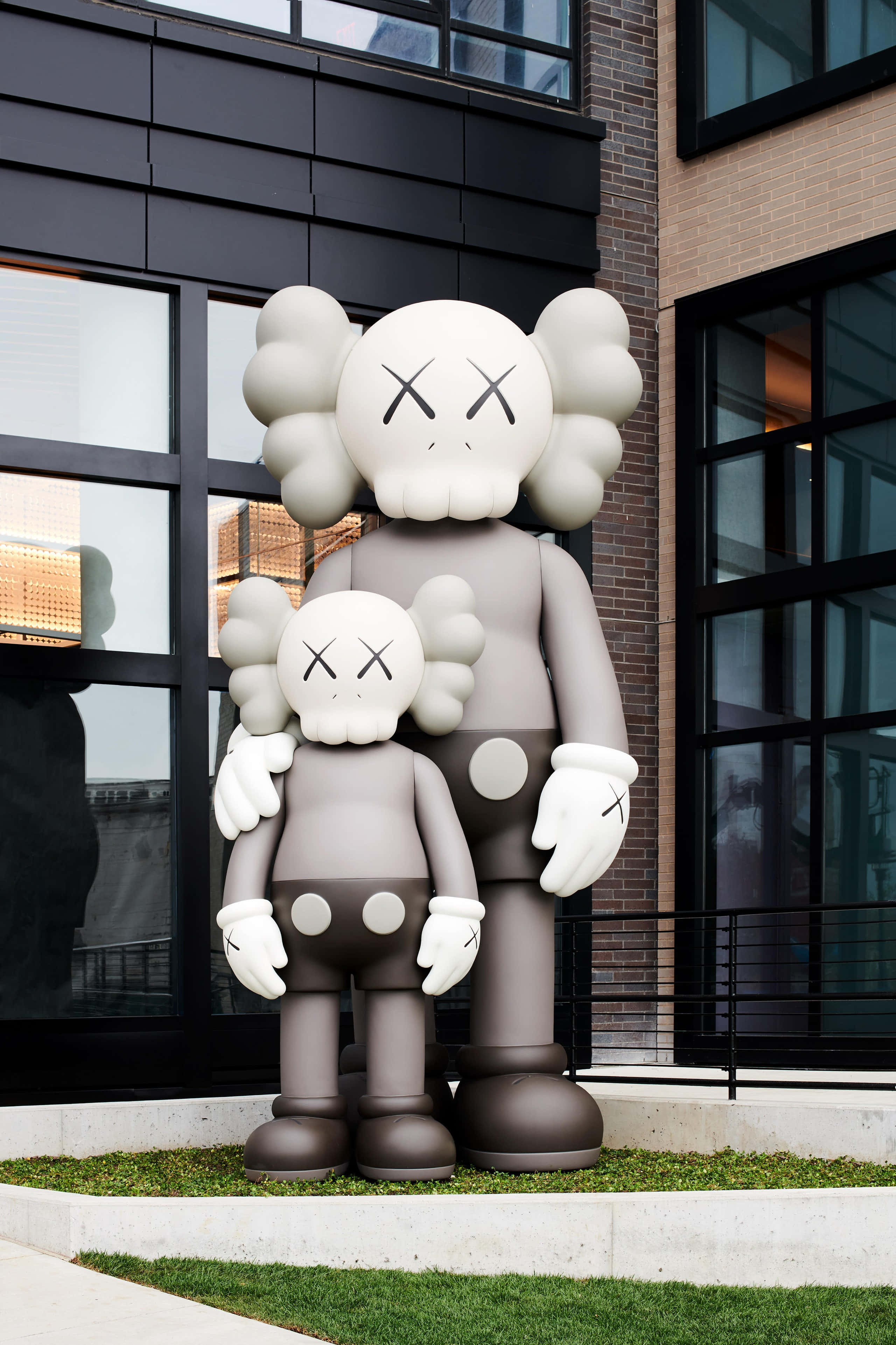 Kaws 推出全新雕塑系列 壁紙 — 兩個巨大的灰色KAWS Companion雕像並排站在現代磚和玻璃建築外牆前的草地上。