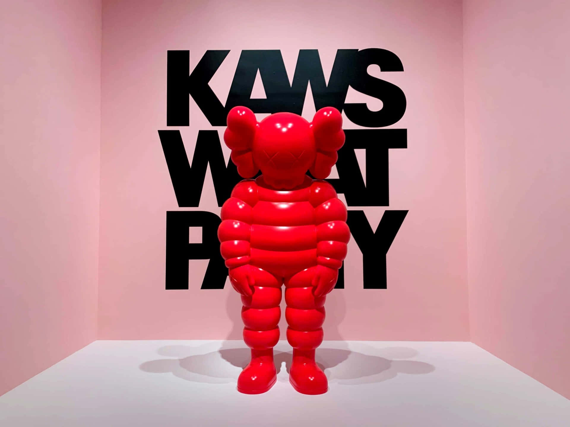 KAWS 為合作精神喝采 壁紙 — 一個光澤的紅色KAWS Companion雕塑放在白色底座上，背景是粉紅色的牆壁，上面有粗體黑色'KAWS WHAT PARTY'文字。