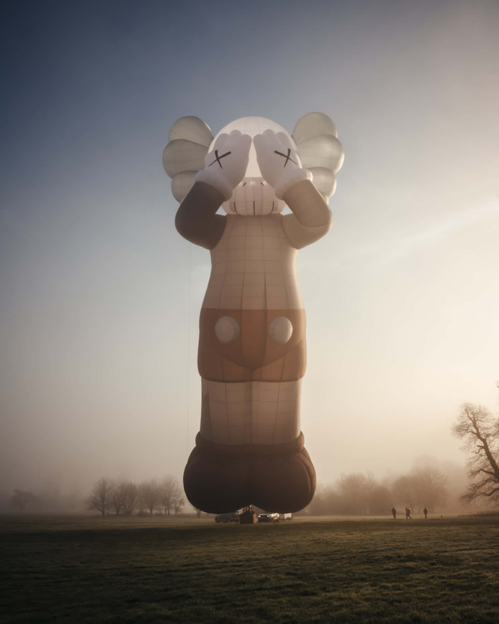 KAWS 的鮮豔多彩世界 壁紙 — 一個巨大的KAWS氣球雕塑在日出時分矗立於霧氣瀰漫的田野中，遠處有人們在觀望。