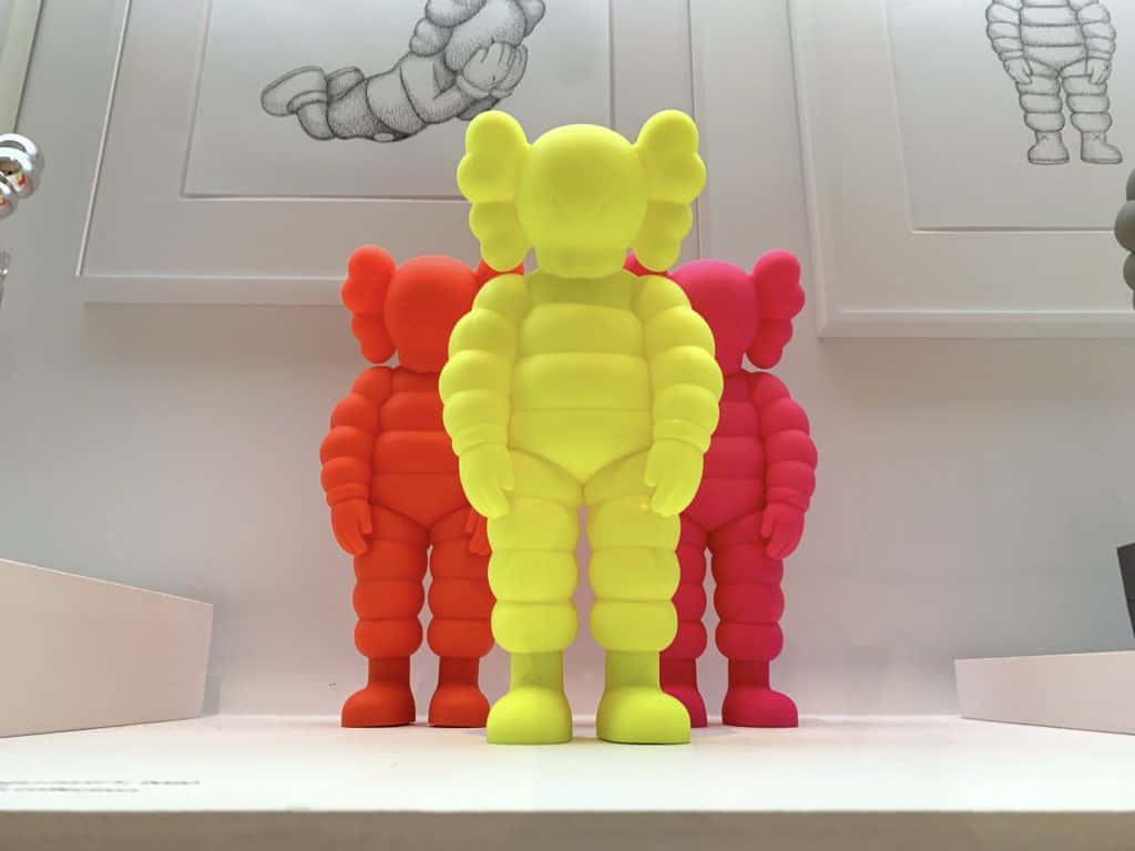 KAWS 藝術作品提供獨特視角 壁紙 — 三個色彩鮮豔的KAWS毛絨公仔，黃色、橙色和粉紅色，排成一排放在白色架子上，背景牆上有框著的素描。