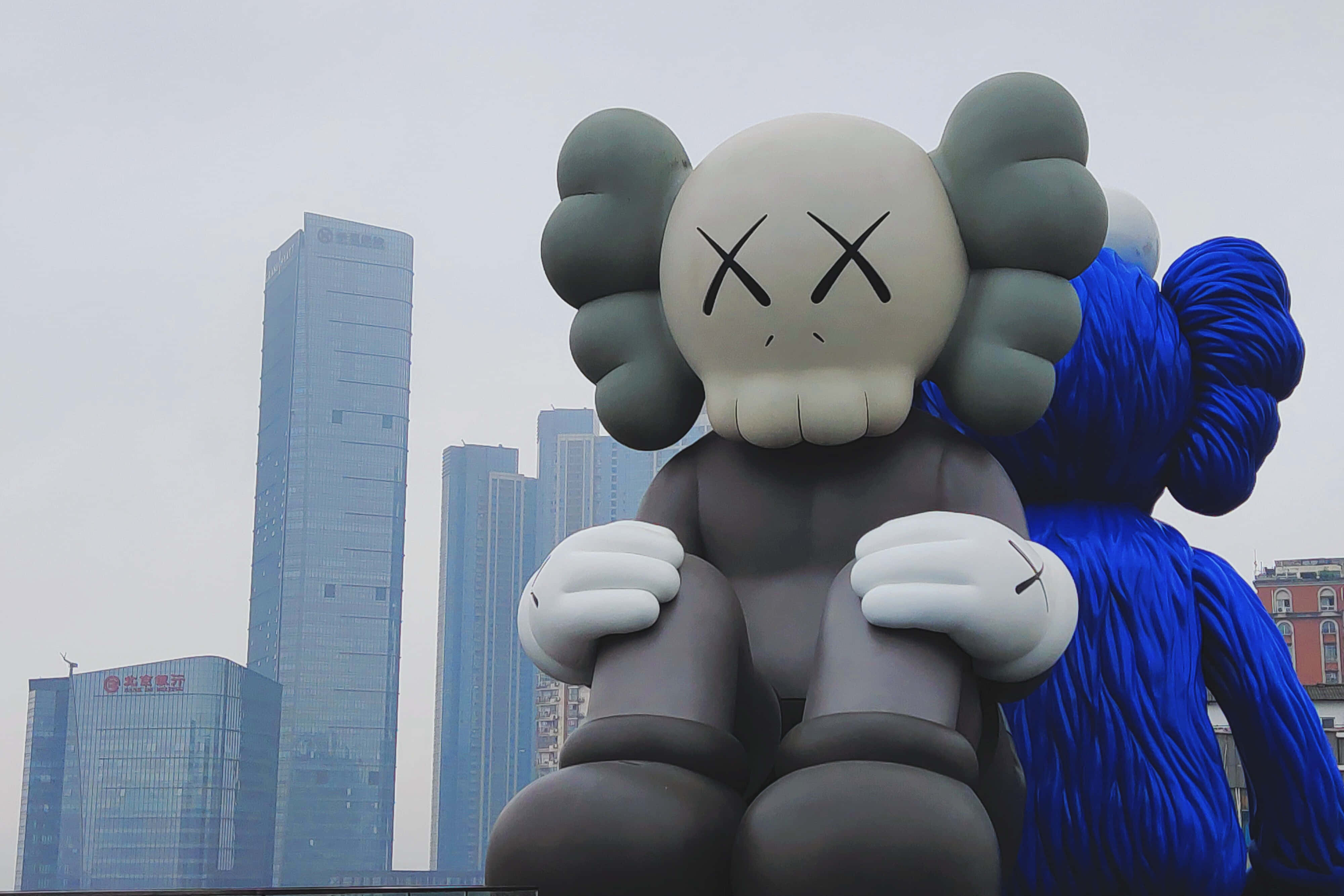 KAWS：街頭藝術家、文化象徵與設計師 壁紙 — 一個巨大的灰色KAWS雕塑，帶有X形眼睛，在陰天背景下，現代玻璃摩天大樓前靜靜地坐著。