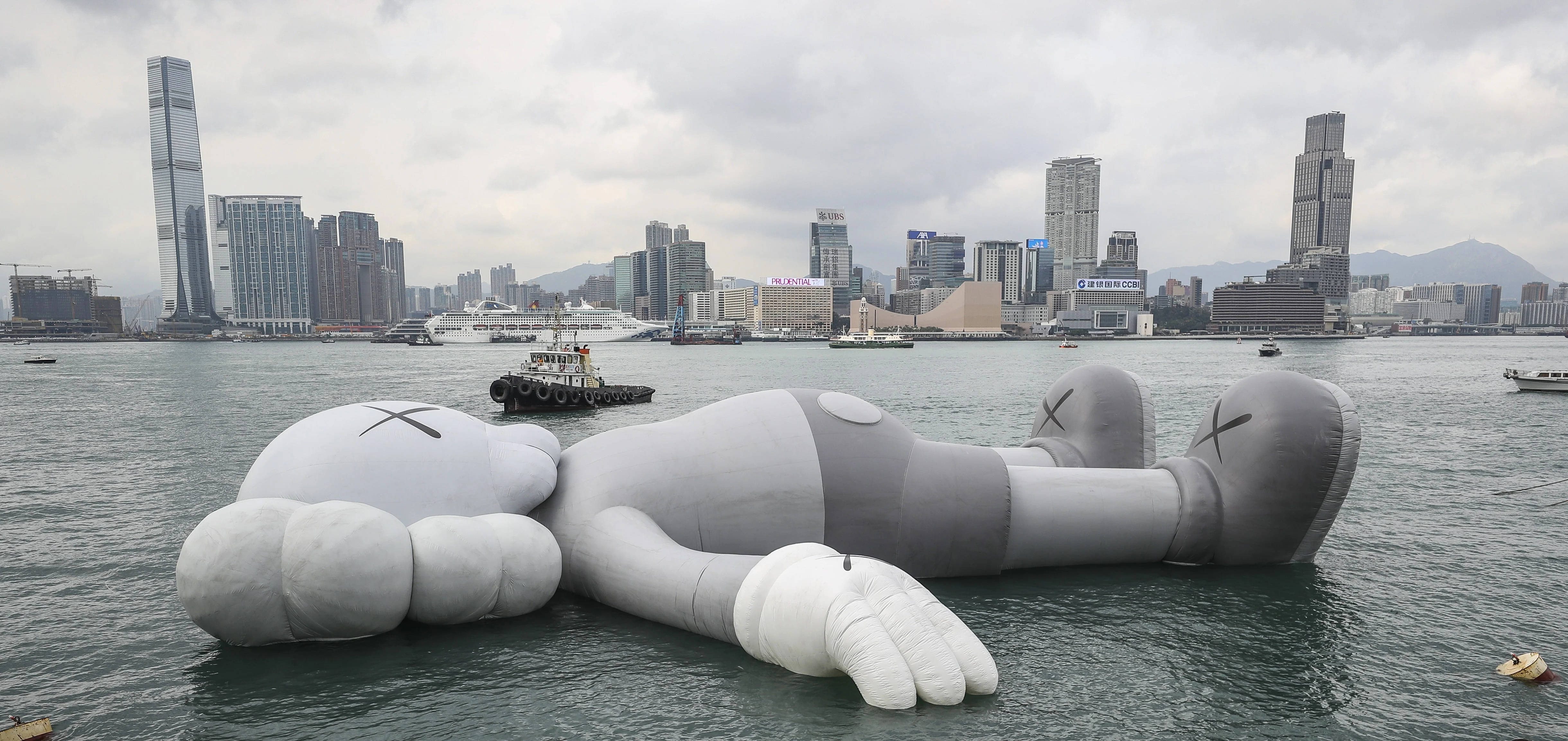 KAWS 香港海港 壁紙 — 一個巨大的灰色充氣KAWS雕塑在陰天背景下，在港口水中寧靜地漂浮，背後是密集的現代城市天際線。