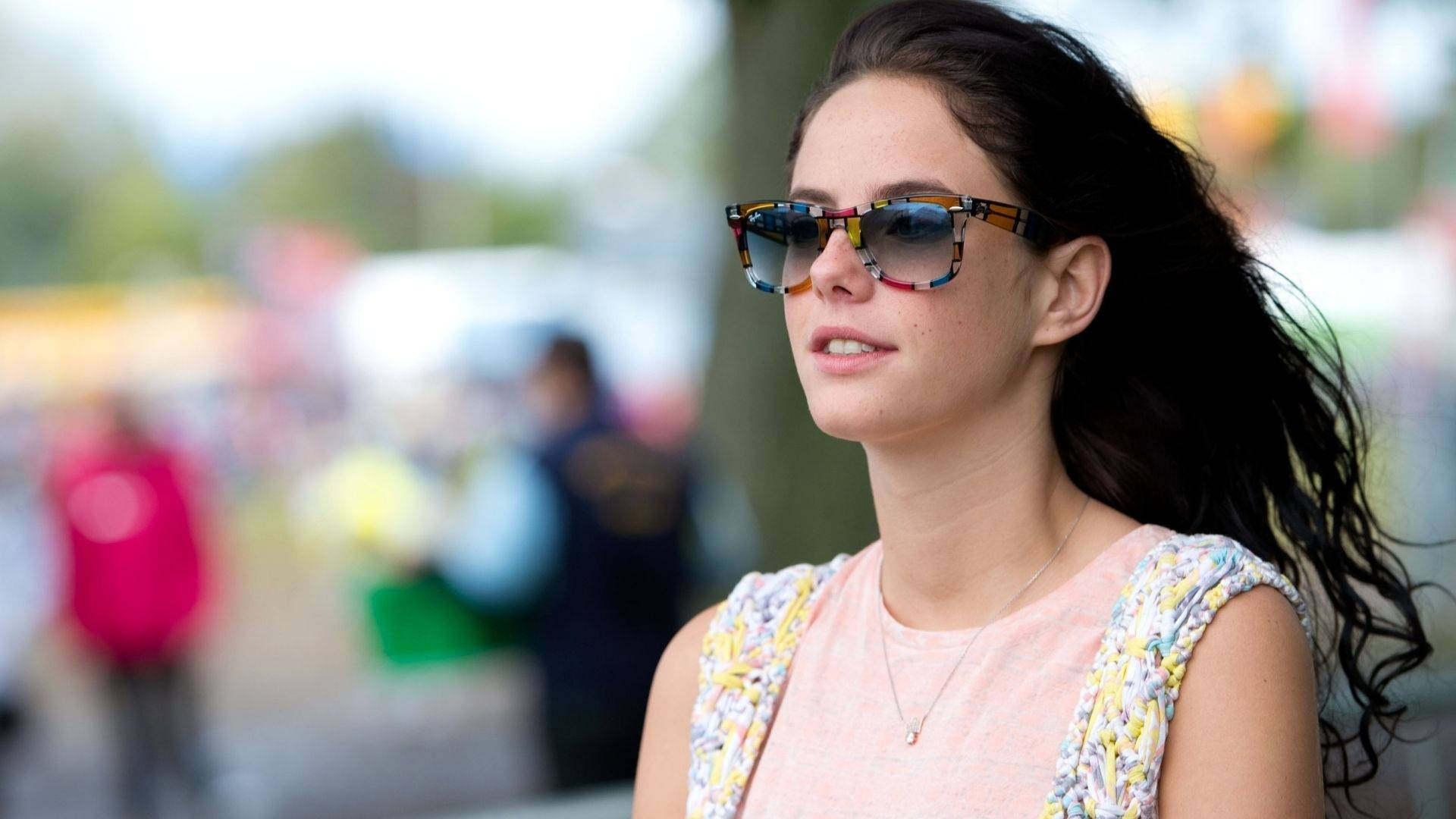 Kaya Scodelario 夏日風情壁紙 壁紙 — 一位深色頭髮、戴著圖案太陽眼鏡的女性，在陽光明媚的戶外環境中若有所思地向上凝視。