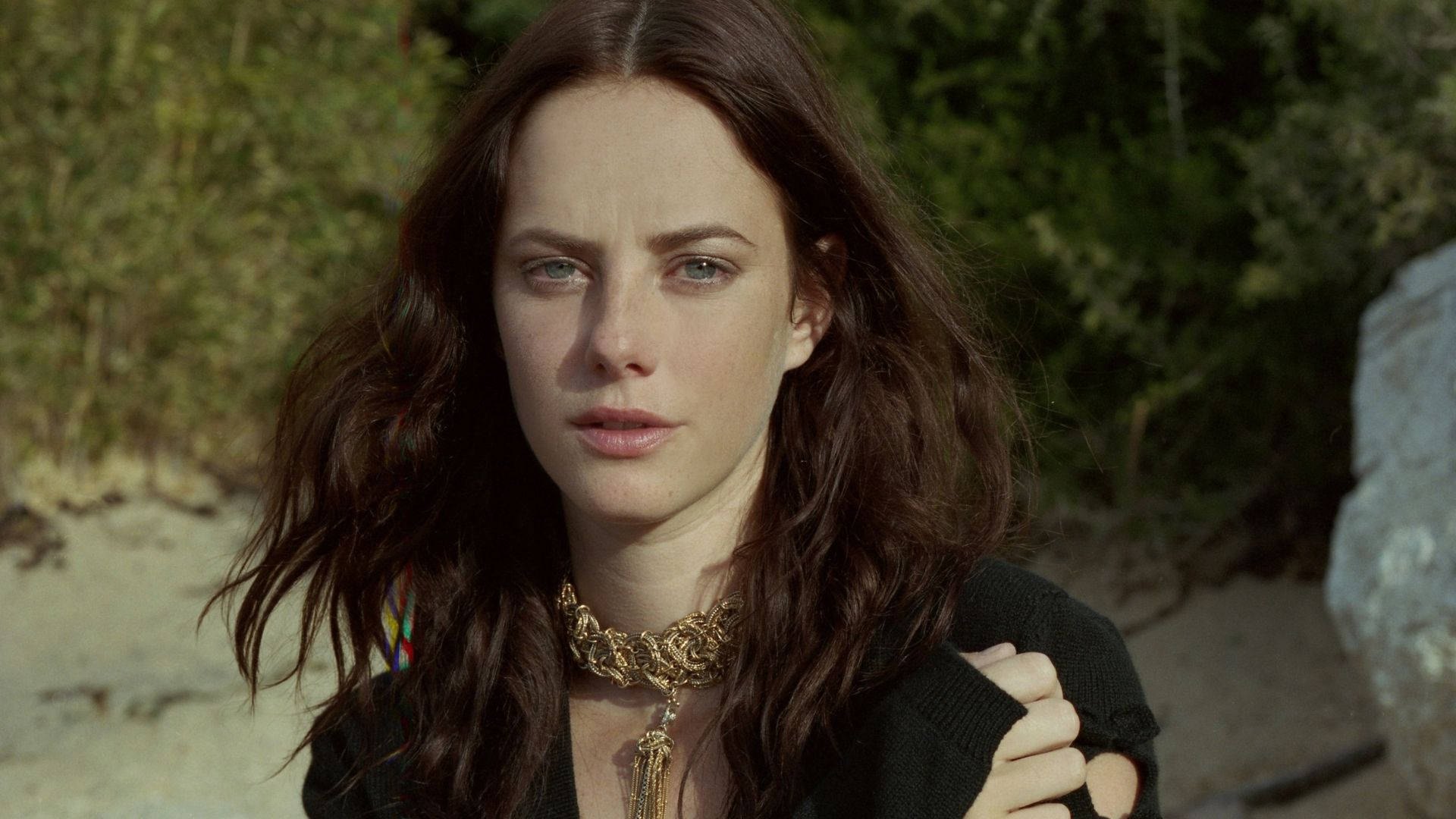 Kaya Scodelario 露天陽光壁紙 壁紙 — 一位擁有長黑髮和金色頸鏈的女性，在模糊的自然背景下專注地凝視。