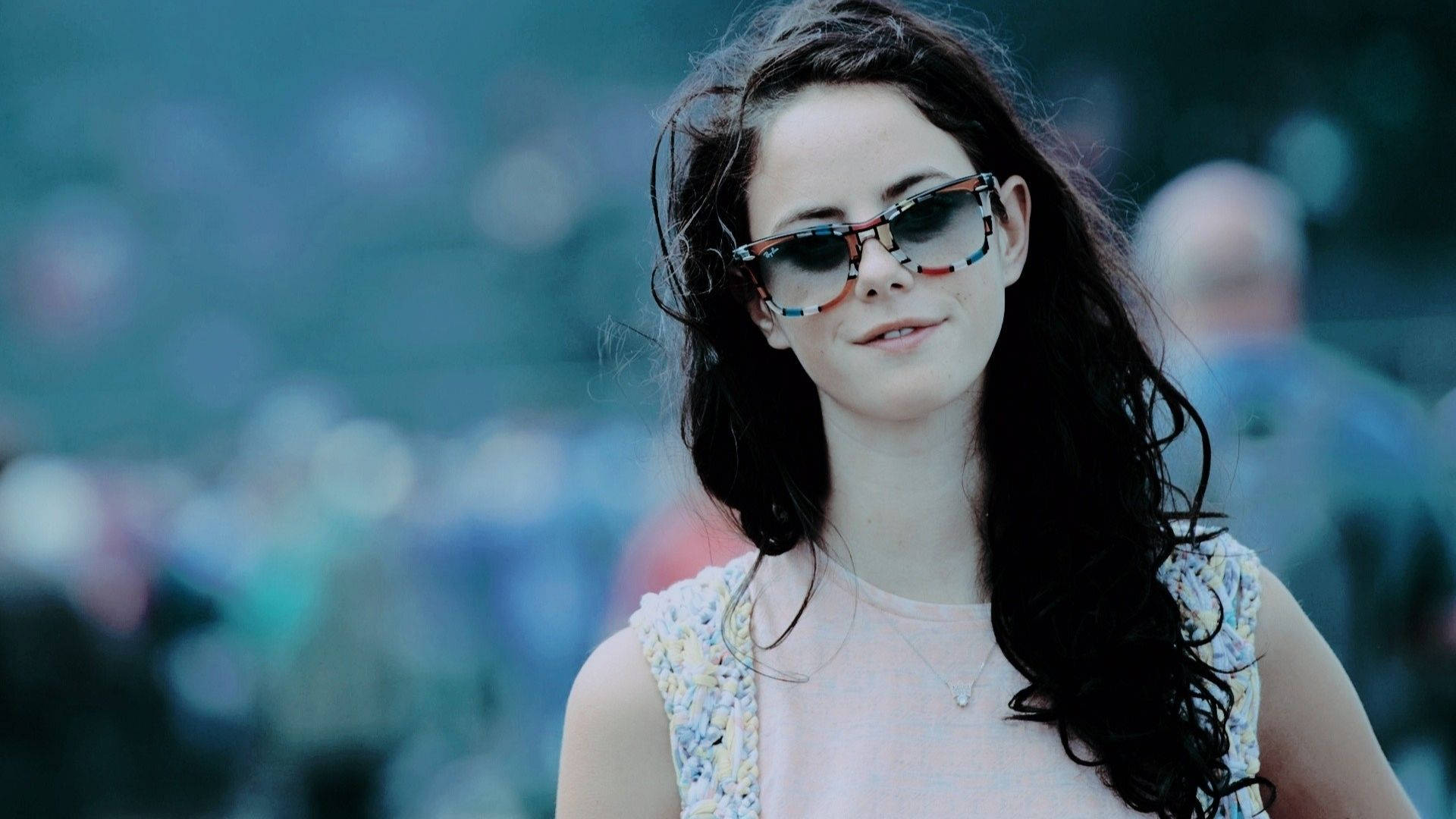 Kaya Scodelario 深色妝容壁紙 壁紙 — 一位年輕女性，深色波浪長髮，戴著彩色太陽眼鏡，在模糊的人群背景中溫柔微笑。