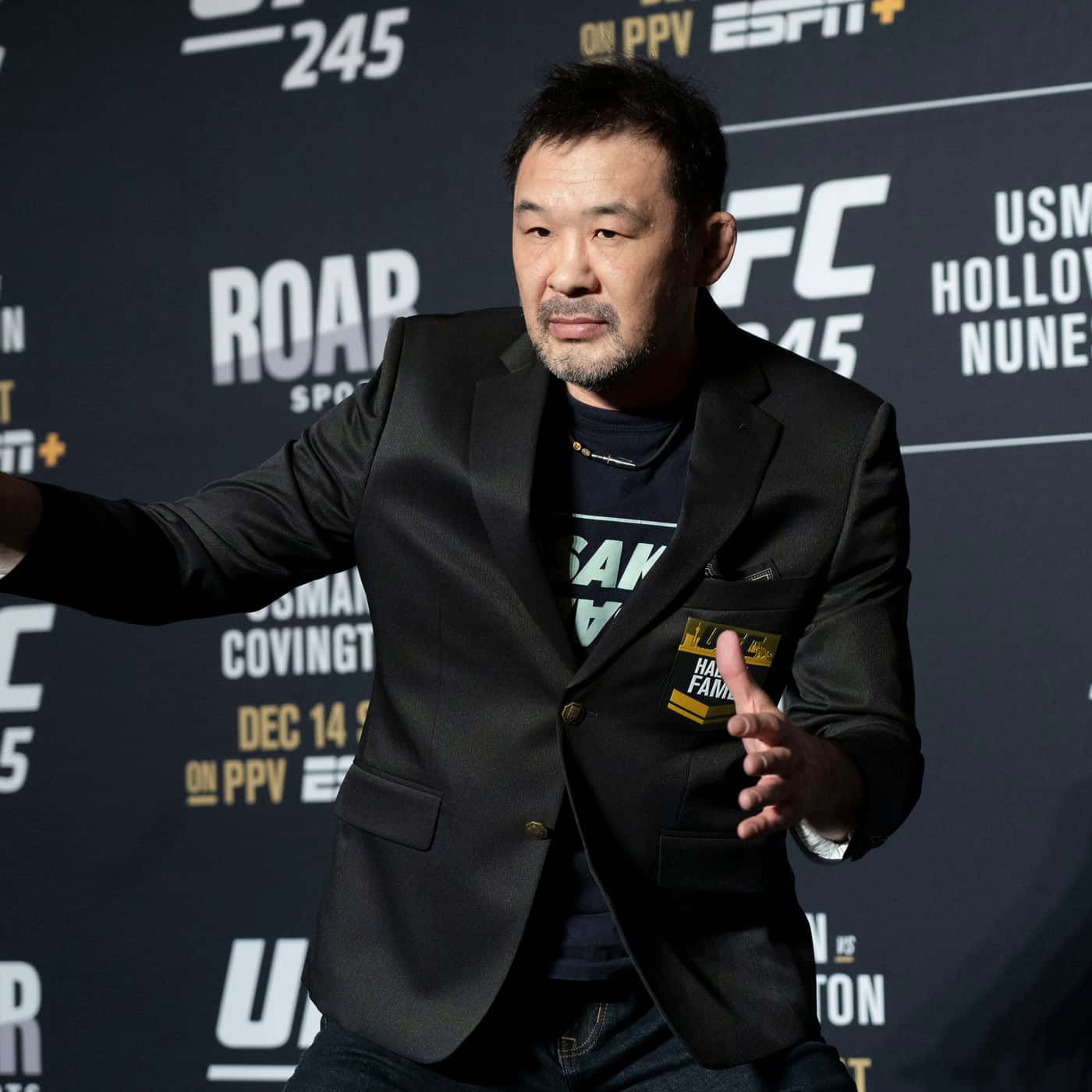 Kazushi Sakuraba出席UFC 245媒體日 壁紙 — 一名身穿黑色西裝外套的男子在UFC記者會上自信地做手勢，身後有宣傳橫幅。