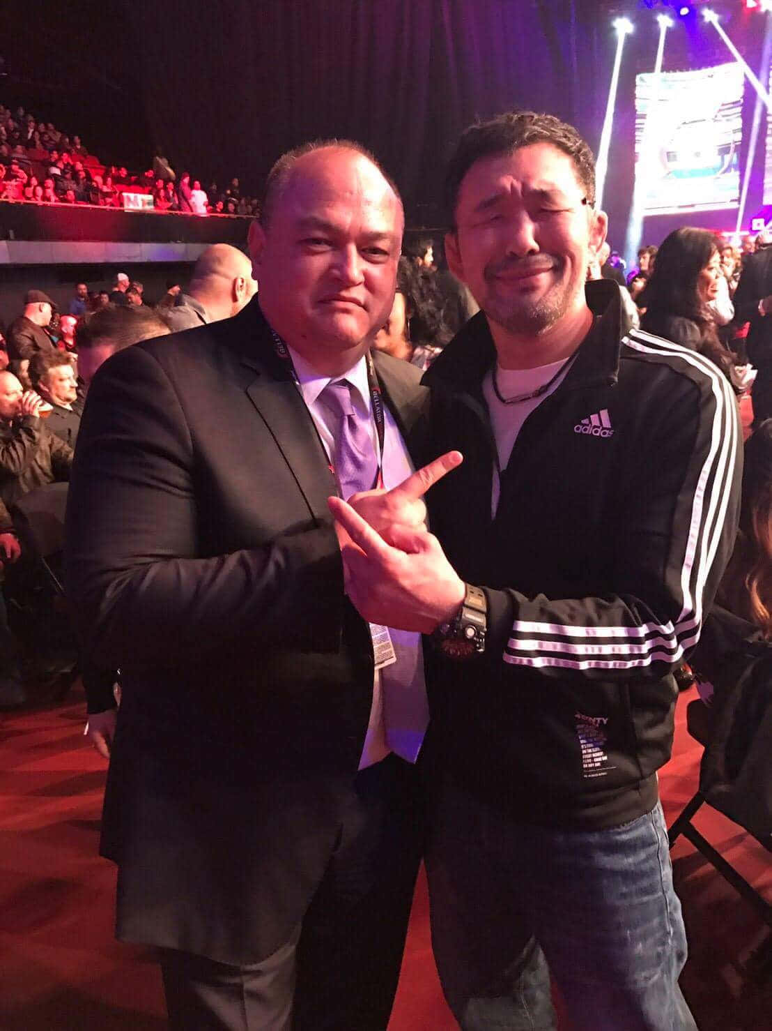 Kazushi Sakuraba 出席活動 壁紙 — 兩名男子在擠滿人的活動中，在舞台燈光下拍照，身後有坐著的觀眾。