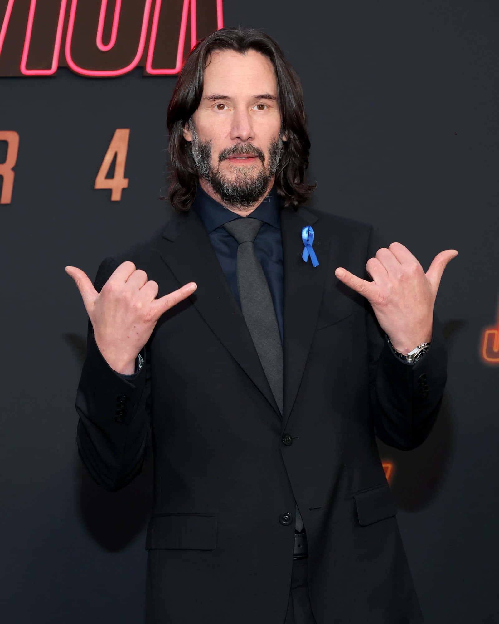 Keanu Reeves 桌布 壁紙 — 奇洛李維斯身穿黑色西裝，在電影首映禮上佩戴藍色絲帶別針，做出沙卡手勢。