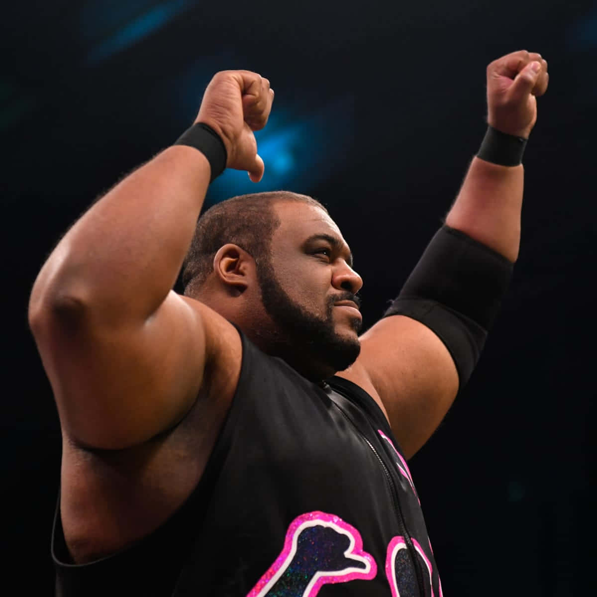 Keith Lee AEW比賽勝利慶祝 壁紙 — 一位肌肉發達的摔角手穿著帶有粉紅色閃粉標誌的黑色背心，在明亮的舞台燈光下勝利地高舉雙拳。