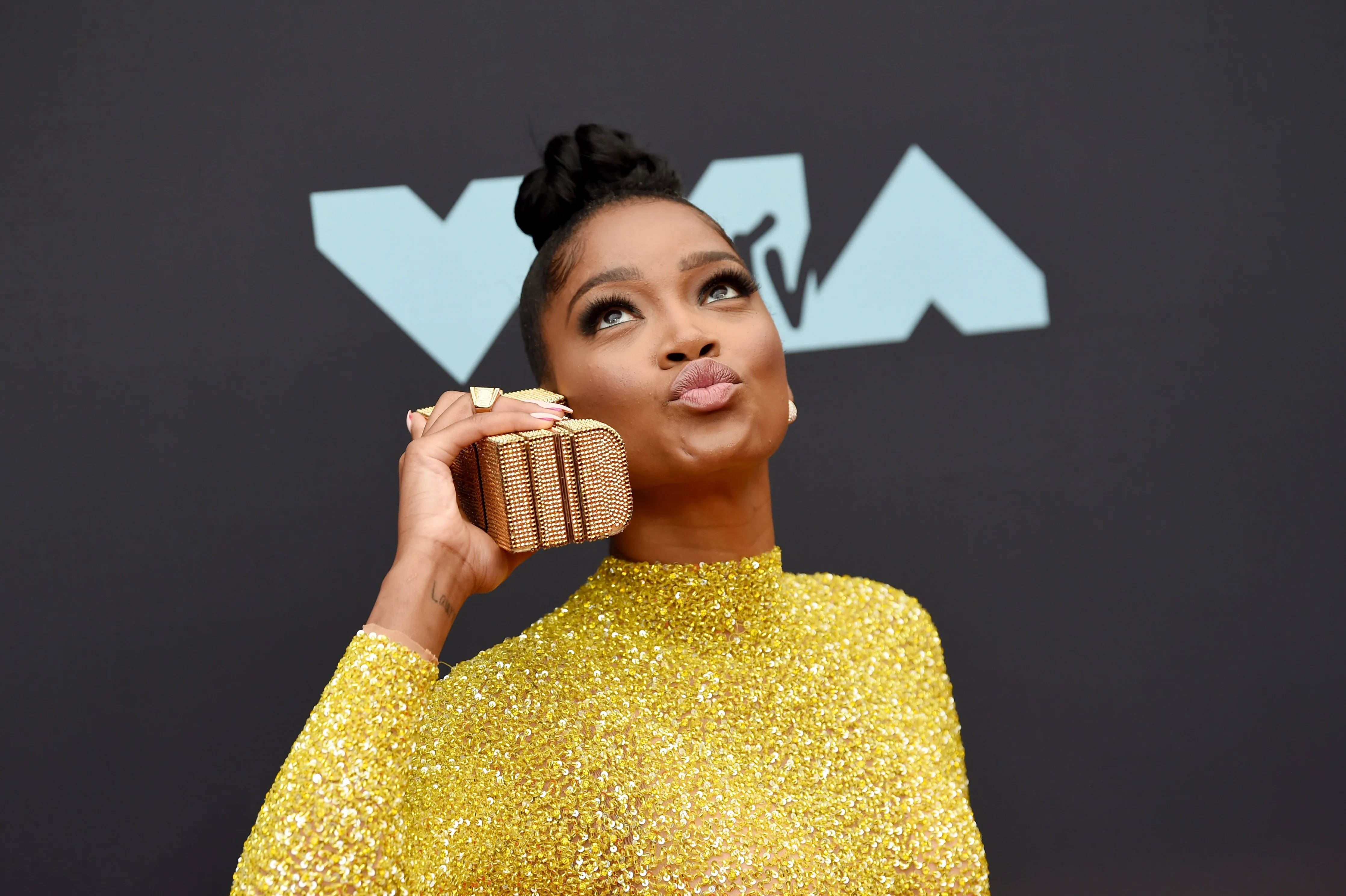 Keke Palmer 2019 VMA 壁紙 — 一位身穿閃亮金色連衣裙的女性在紅地毯活動中俏皮地親吻一個鑲有寶石的手拿包。