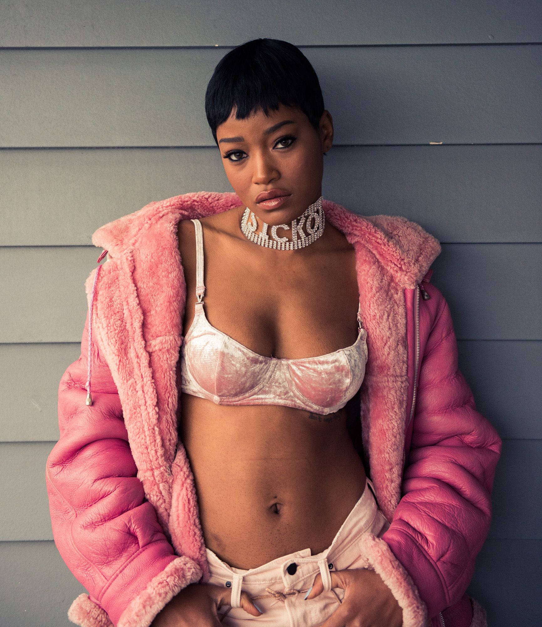 Keke Palmer Coveteur 雜誌拍攝 壁紙 — 一位自信的短黑髮女性穿著粉紅色絨毛外套，內搭白色胸罩和牛仔褲，靠著灰色牆壁。