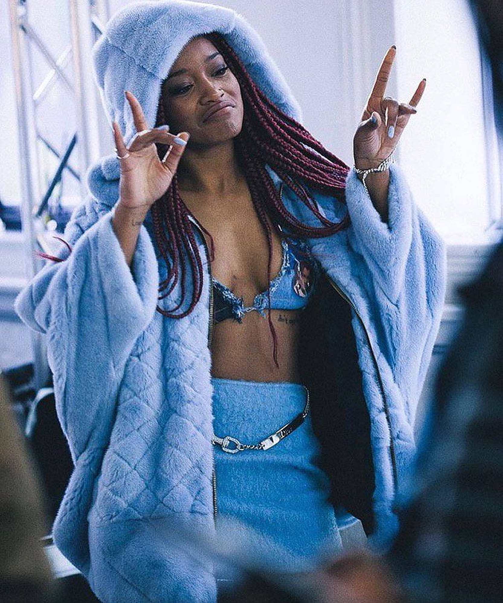 Keke Palmer 美學攝影 壁紙 — 一位穿著蓬鬆淺藍色連帽外套的女性，在穿著牛仔布內衣和配套裙子的同時做出搖滾手勢。