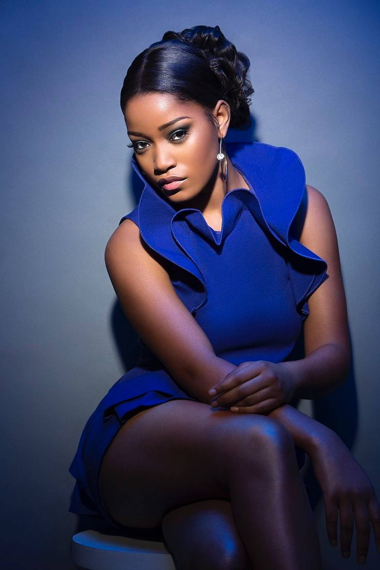 Keke Palmer 藍色禮服 壁紙 — 一位沉穩的女性身穿大膽的藍色連衣裙，在冷色調的攝影棚背景中，配合戲劇性的燈光，自信地坐著。