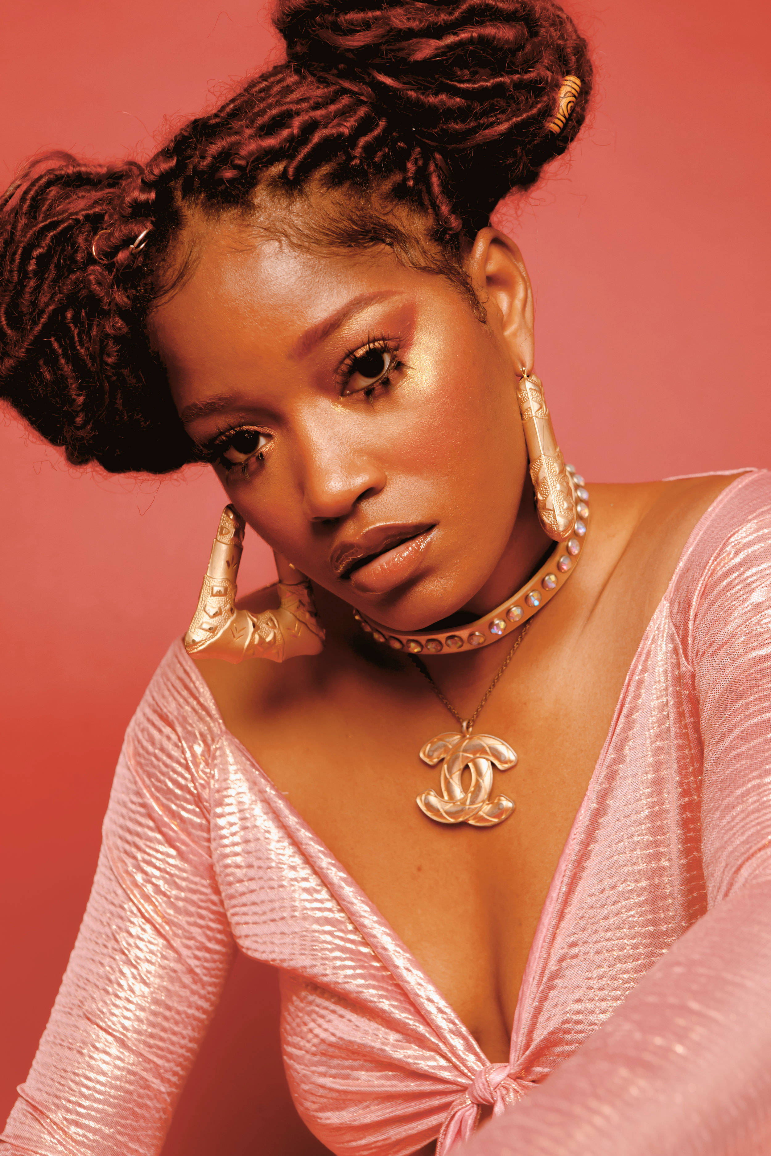 Keke Palmer Lula Hyers 近距離特寫 壁紙 — 一位自信的女性，辮子頭髮和金色珠寶，在溫暖的珊瑚色背景前擺姿勢，穿著閃亮的粉紅色上衣。