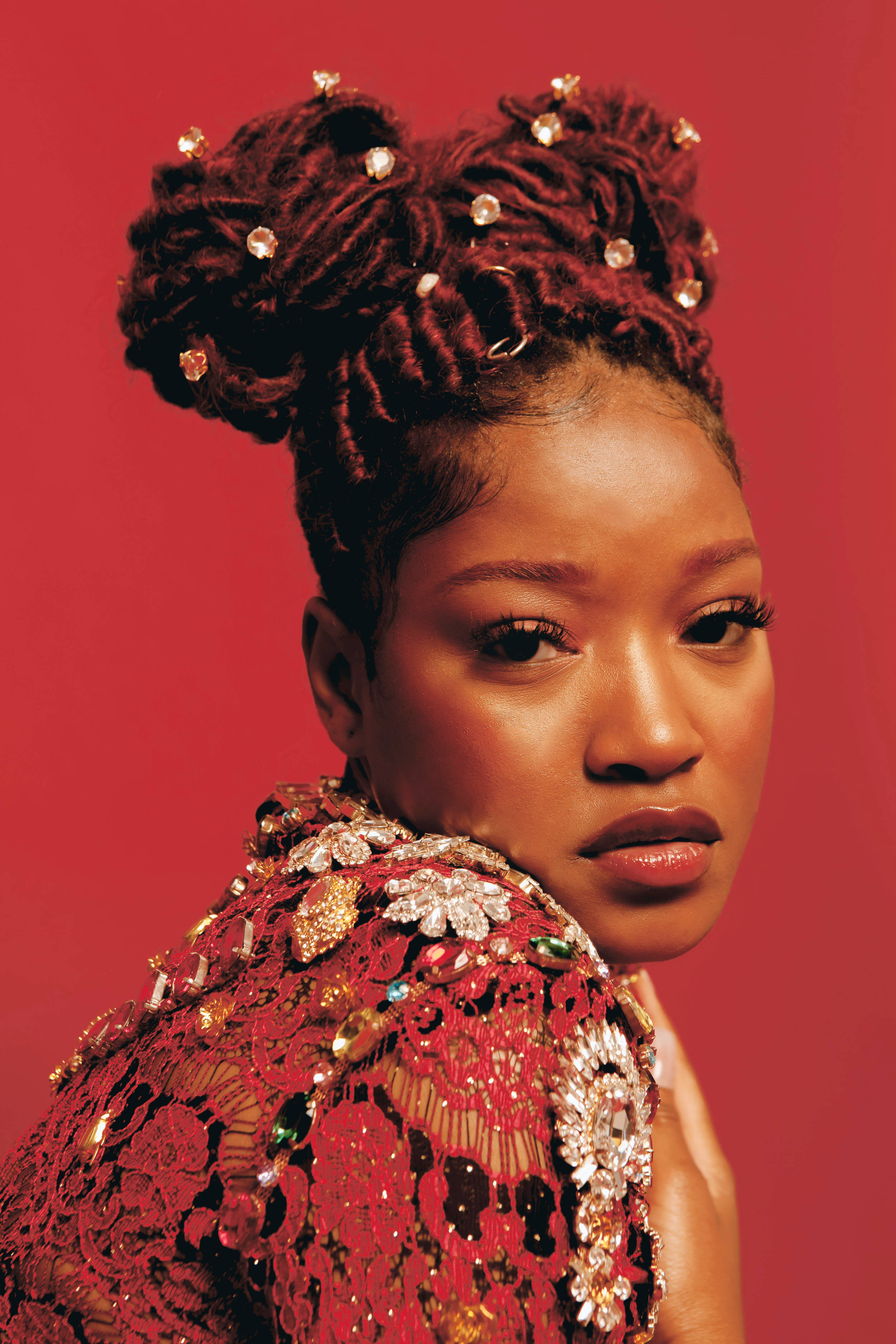 Keke Palmer Lula Hyers 紅色系列 壁紙 — 一位女士梳著精緻的紅色辮子盤髮，飾有珠寶，在純紅色背景前溫柔凝視。