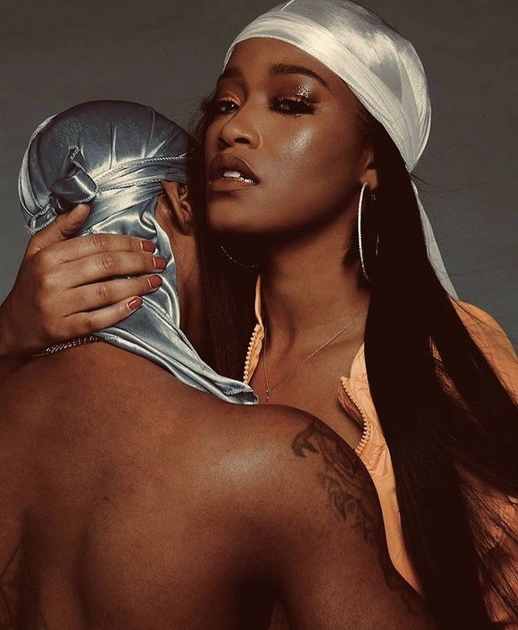 Keke Palmer 复古風情 壁紙 — 一位戴著白色緞面頭巾的女性，在柔和的灰色背景前擁抱一位戴著銀色緞面頭罩的男性。