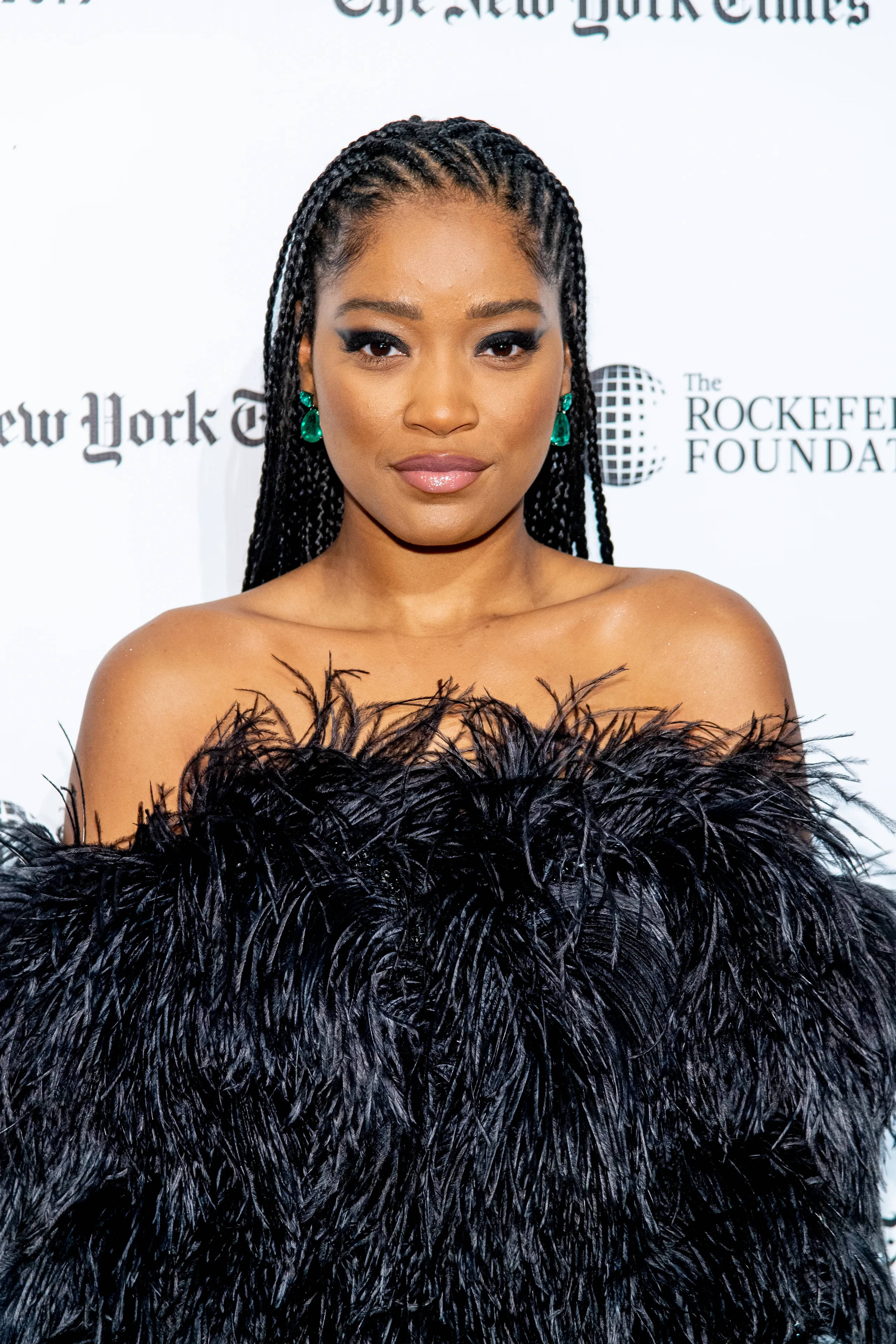 Keke Palmer 紐約時報活動 壁紙 — 一位辮子頭髮、戴綠色耳環的優雅女士，在正式場合穿著一件戲劇性的黑色羽毛露肩長裙。