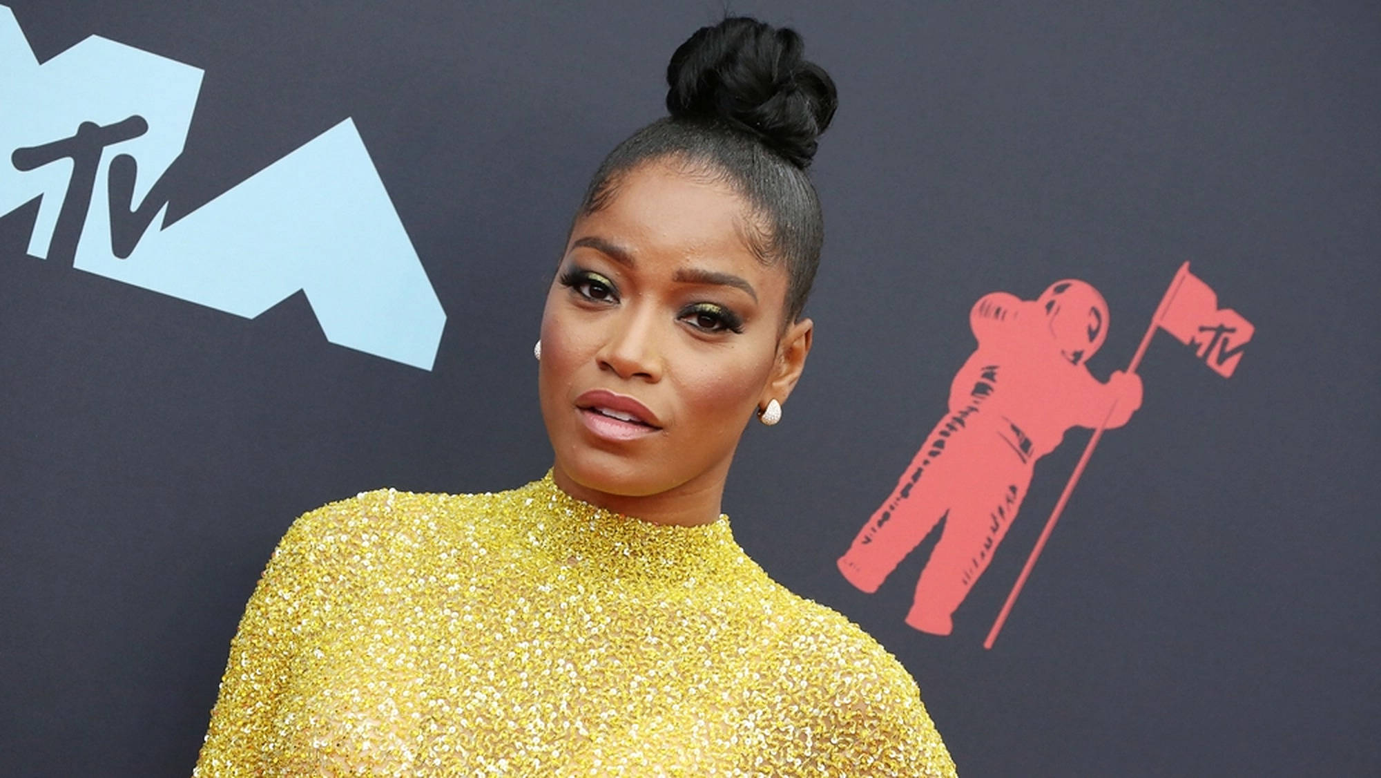 Keke Palmer VMA MTV 壁紙 — 一位身穿閃亮金色連衣裙的女性在帶有MTV標誌和紅色太空人圖案的深色背景前自信地擺姿勢。