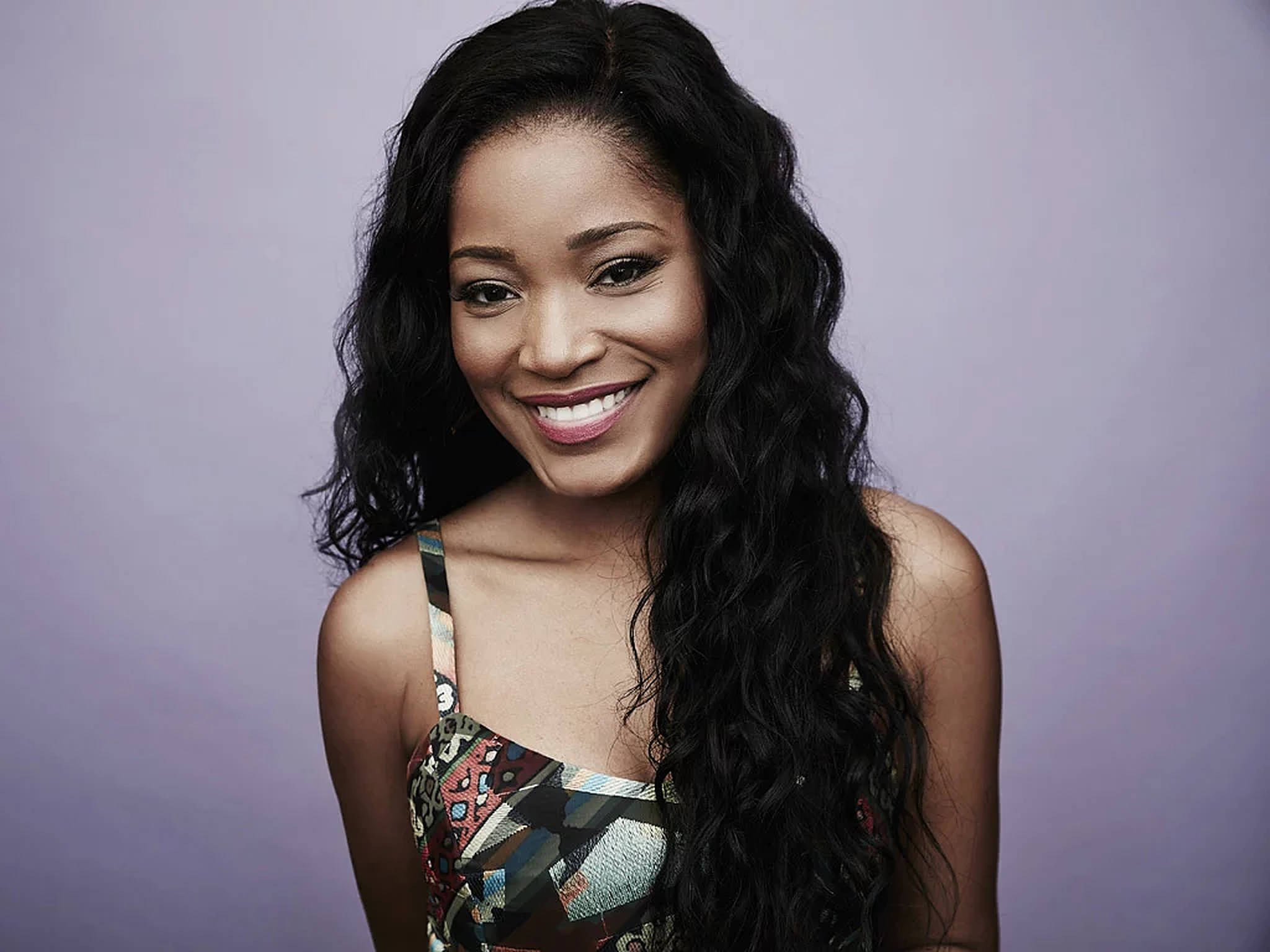 Keke Palmer 卷髮造型 壁紙 — 一位長著深色長捲髮的容光煥發的女性，在柔和的薰衣草色工作室背景下溫暖地微笑。