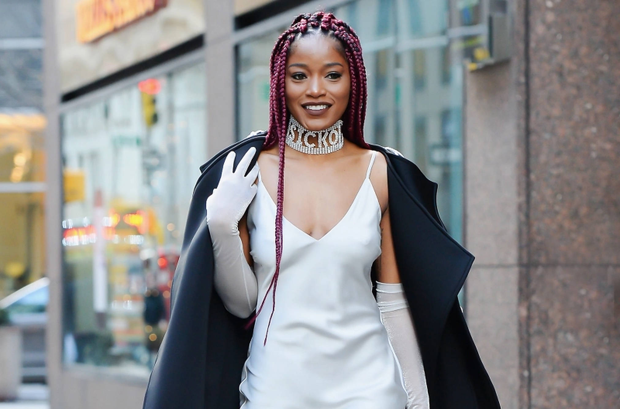 Keke Palmer 白紗禮服 壁紙 — 一位時尚女性，紫色辮子，戴著「STICKY」頸鏈，穿著白色連衣裙和黑色斗篷，在城市街道上微笑揮手。