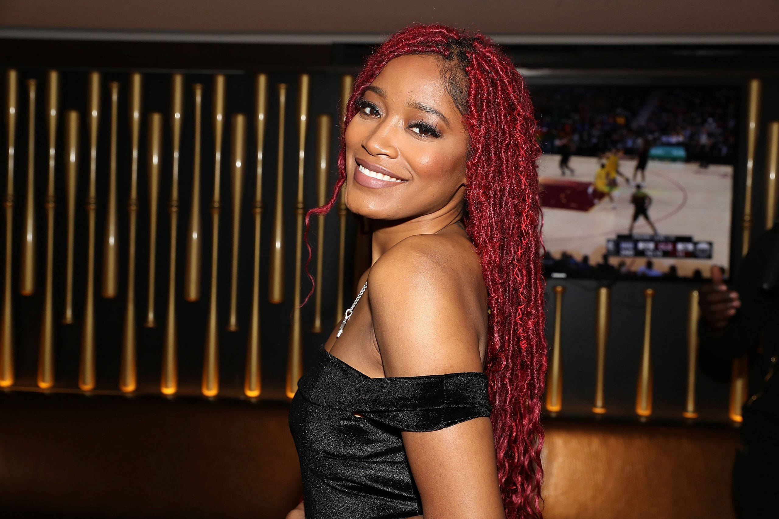 Keke Palmer 紅髮造型 壁紙 — 一位擁有鮮豔紅色辮子的女性，在燈光昏暗的休息室中，身後有金色垂直燈光和螢幕上的籃球比賽，溫暖地微笑。