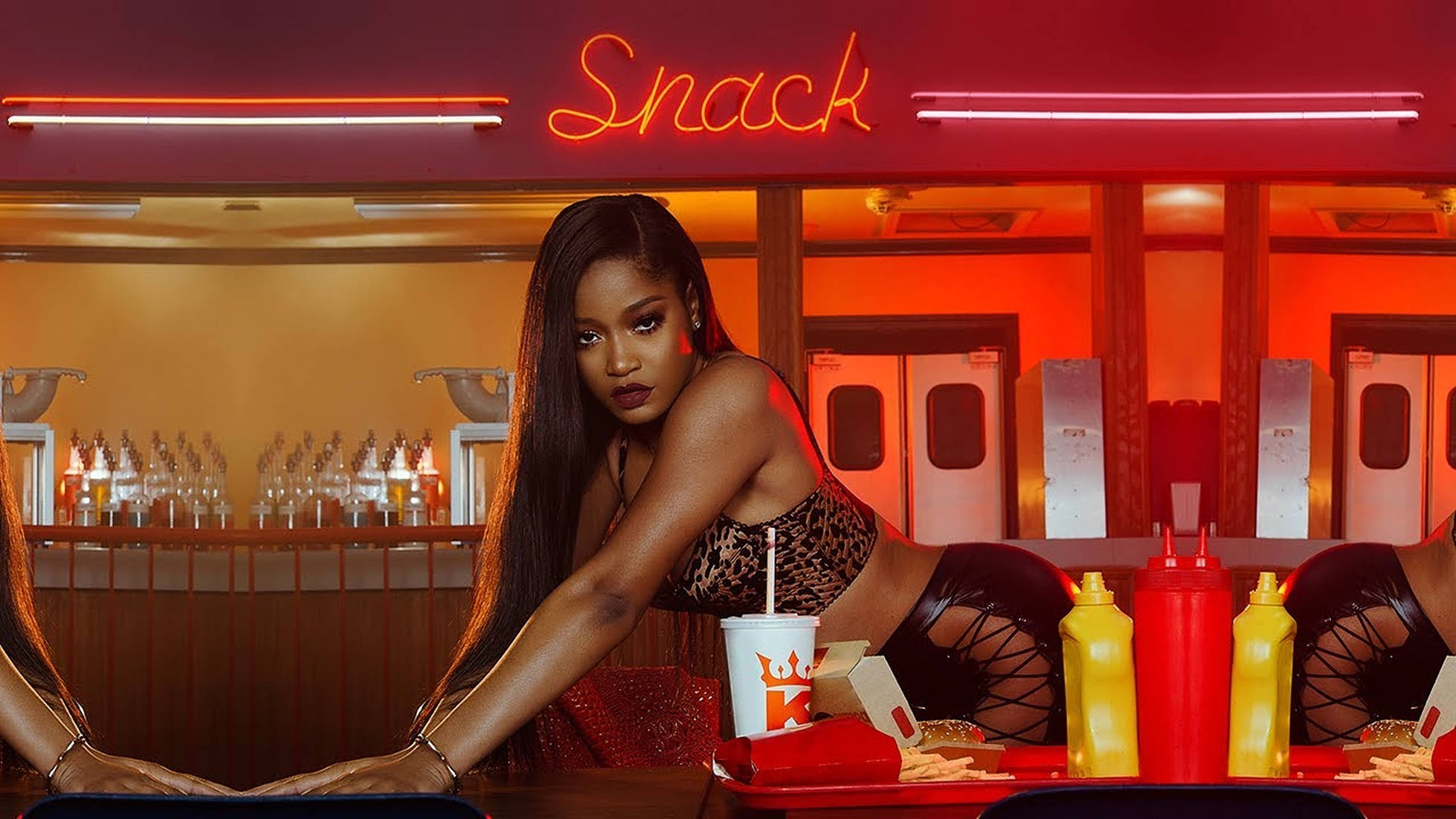 Keke Palmer《Snack》音樂影片 壁紙 — 一名女子在寫著 Snack 的紅色霓虹燈招牌下，俯身靠在小餐館的櫃檯上，周圍放滿調味品和食物托盤。