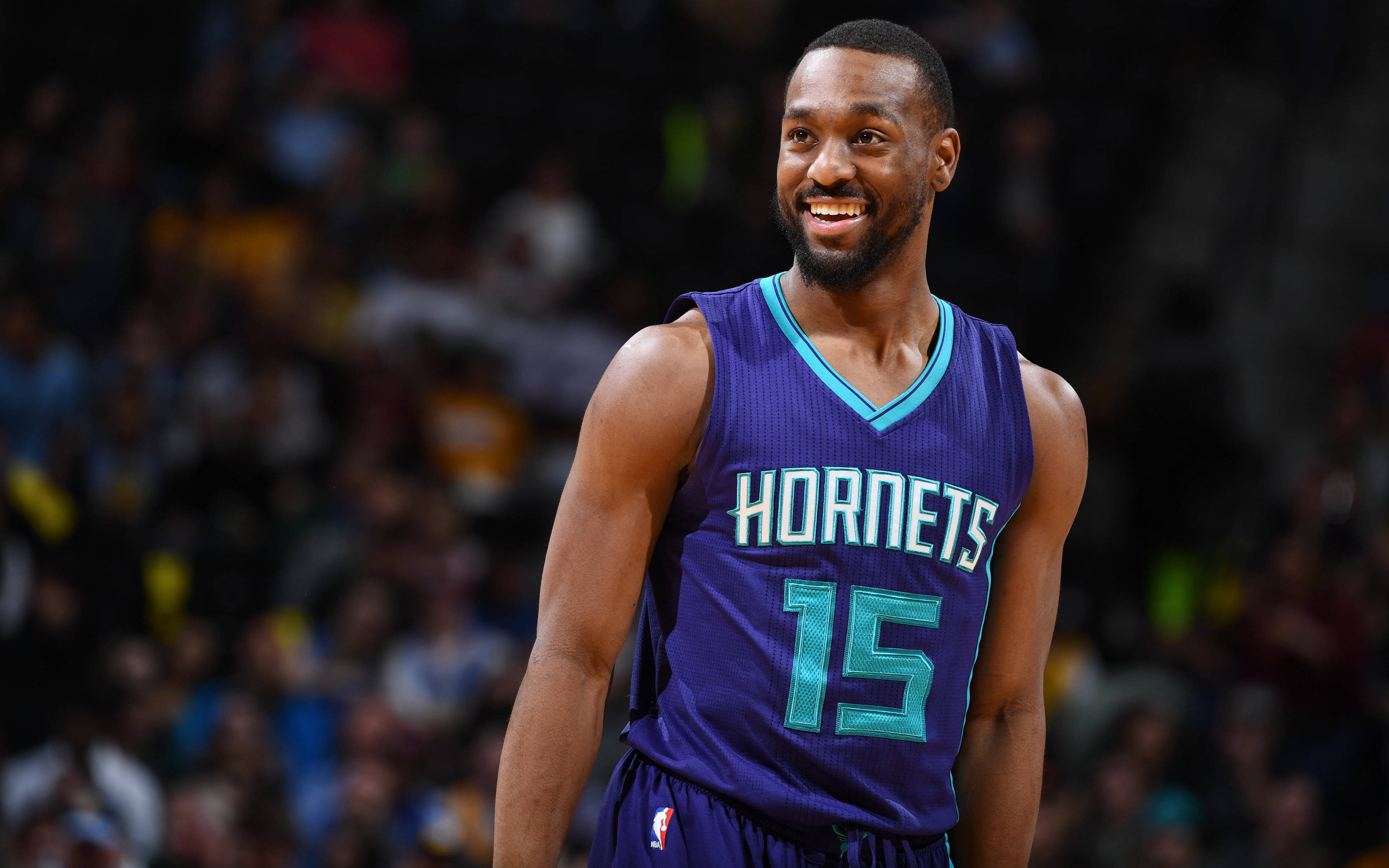 Kemba Walker 微笑 4K 超高清壁紙 壁紙 — 一名微笑的籃球運動員身穿紫色黃蜂隊15號球衣，自信地站在球場上，背景是模糊的觀眾。