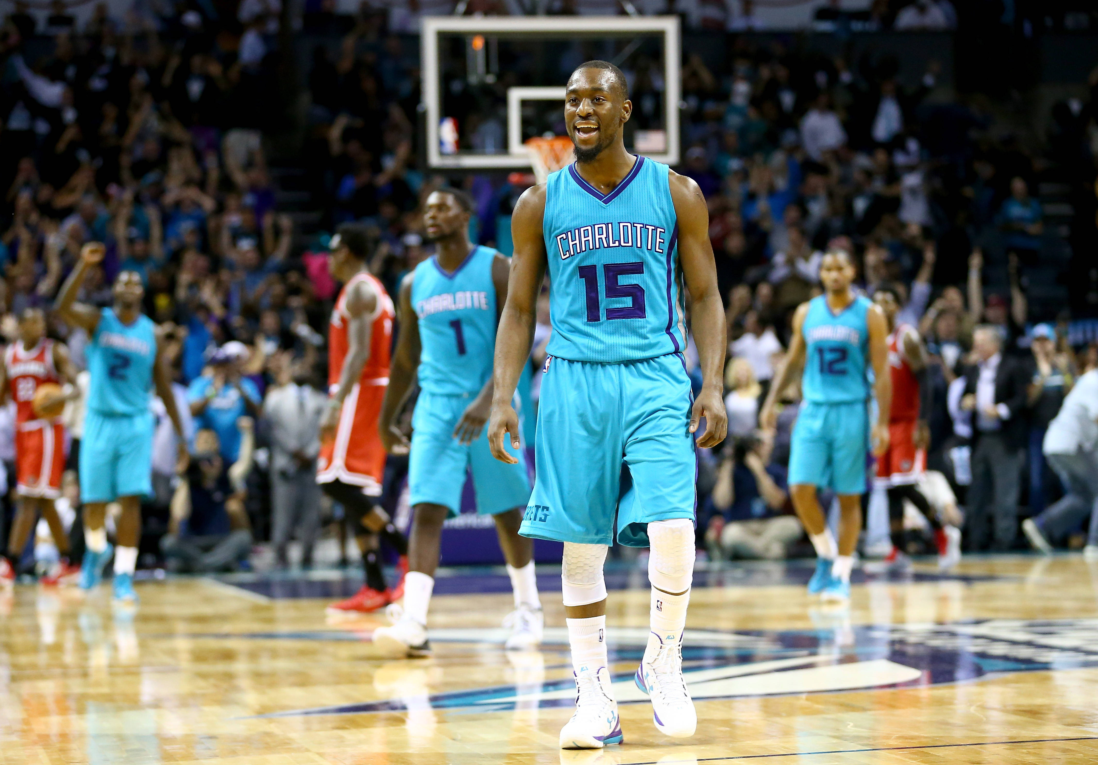 Kemba Walker 黃蜂隊對戰密爾沃基 壁紙 — 一名身穿夏洛特球衣的籃球運動員在光亮的球場上自信微笑，身後有隊友和歡呼的觀眾。