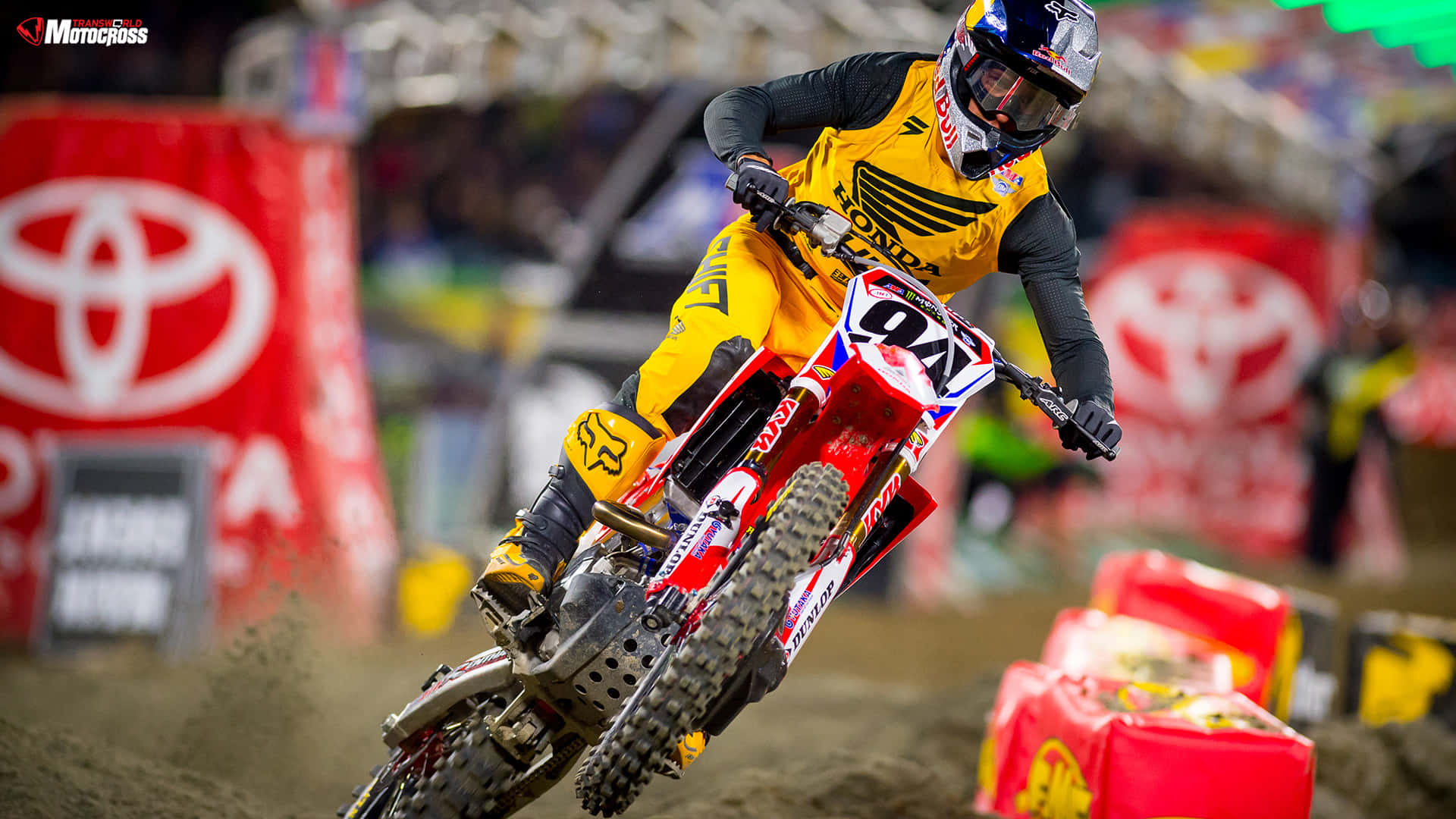 Ken Roczen 展現越野摩托高超技巧 壁紙 — 身穿黃色裝備的越野摩托車手在體育場賽事中，騎著紅色越野車傾斜入彎，濺起泥土。