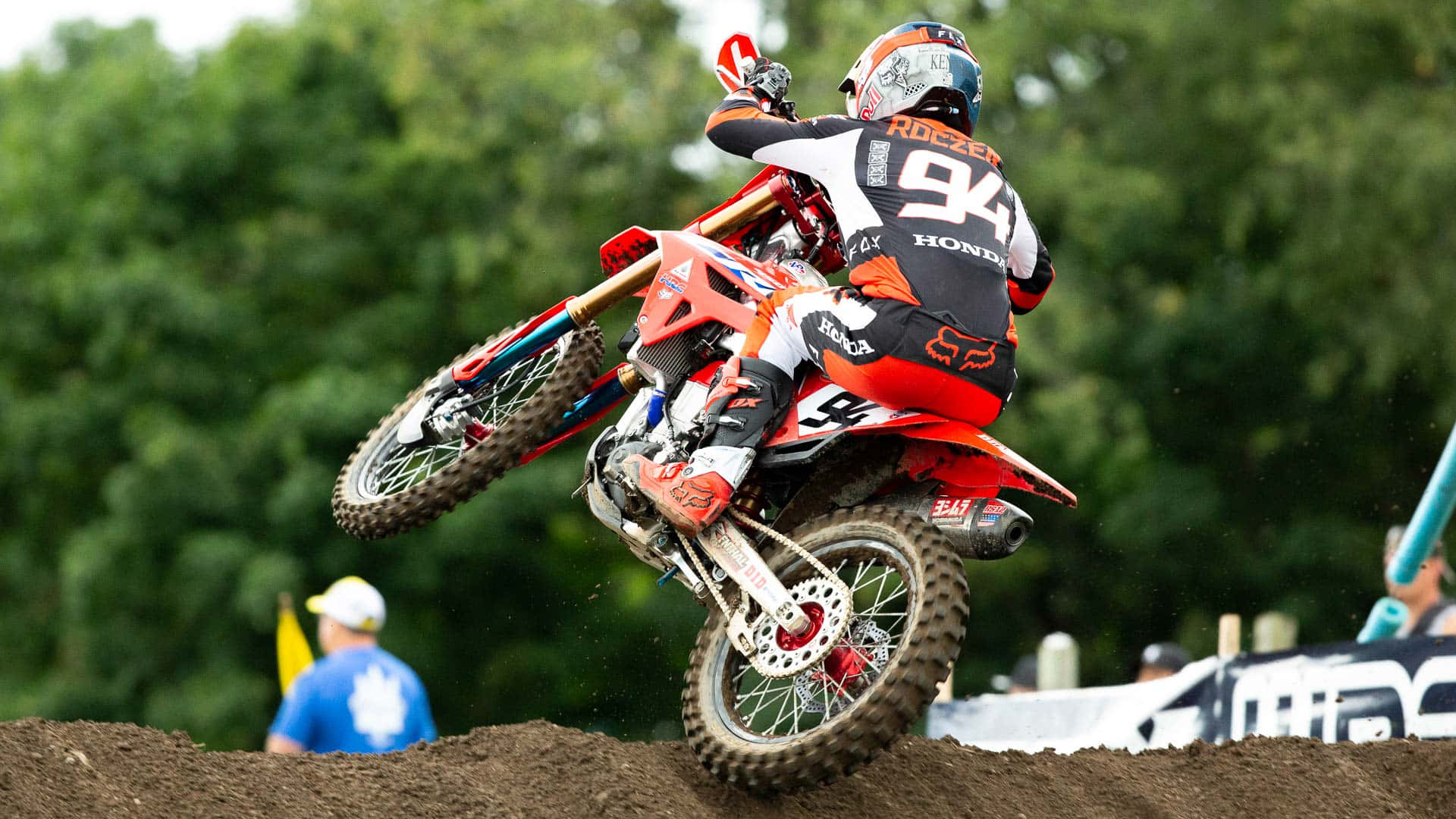 Ken Roczen 攀越賽道山丘桌面 壁紙 — 身穿橙色裝備的越野摩托車手在空中飛翔，騎著紅色本田摩托車越過土坡，背景有綠樹。
