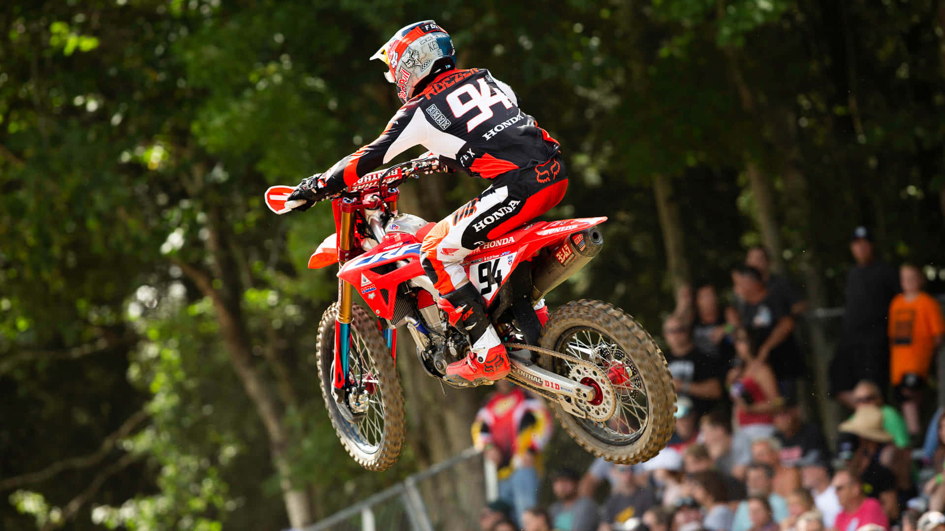 Ken Roczen 紅色飛躍桌面 壁紙 — 一位身穿紅黑裝備的越野摩托車手，騎著紅色本田越野摩托車，在森林賽道上空飛馳，下方有觀眾。