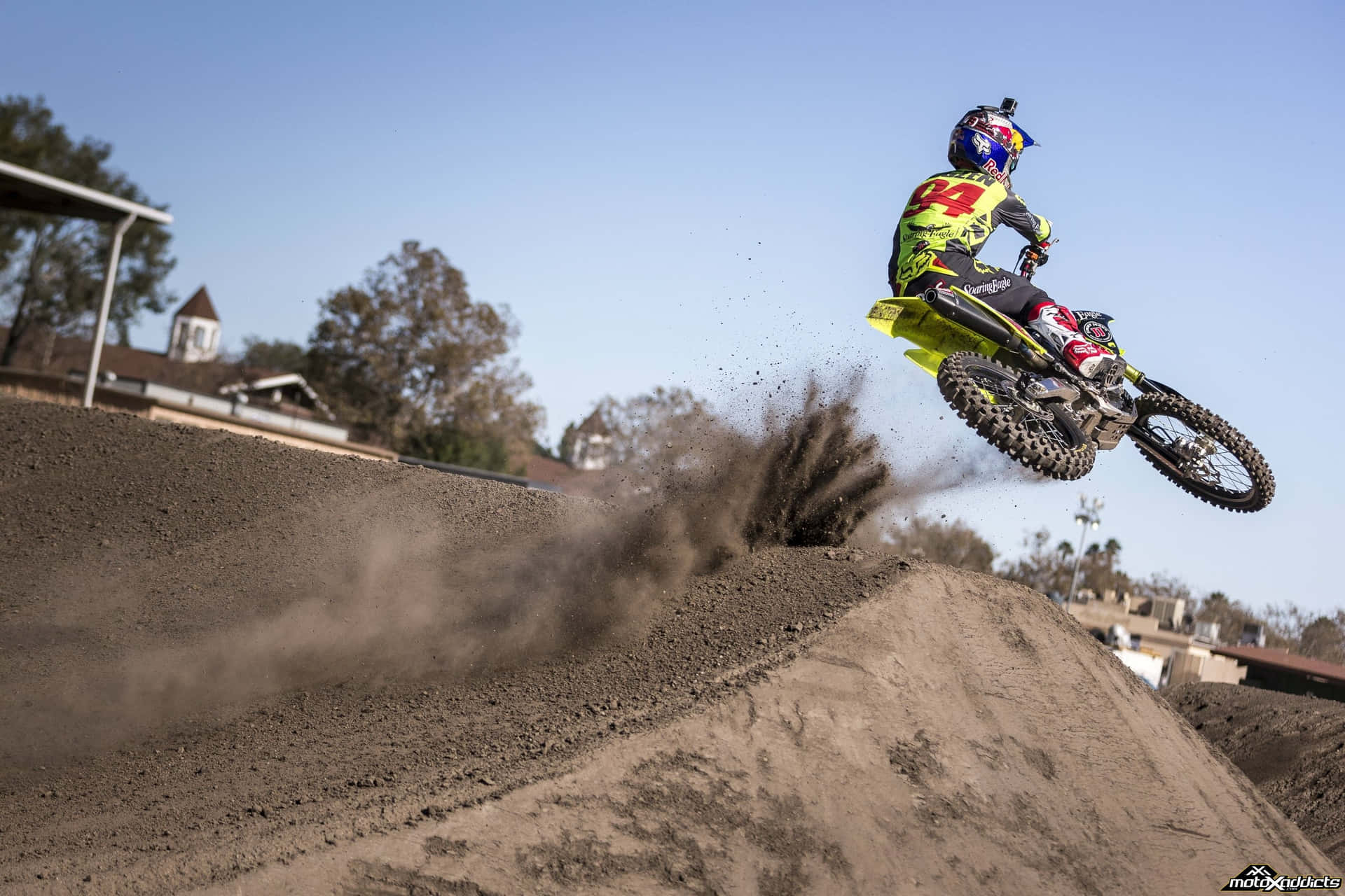 Ken Roczen 自由式越野摩托桌面 壁紙 — 一名身穿鮮豔裝備的越野電單車手飛越泥土跳台，在晴朗藍天下揚起一片塵雲。