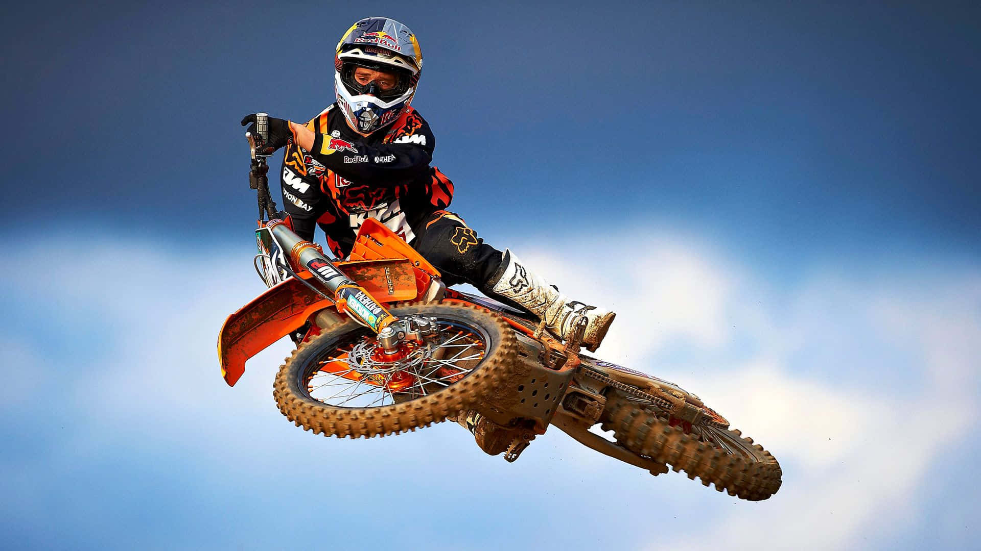 Ken Roczen 越野摩托翻轉桌面 壁紙 — 一名越野摩托車車手騎著橙色越野車在空中飛翔，背景是清澈的藍天和蓬鬆的白雲。