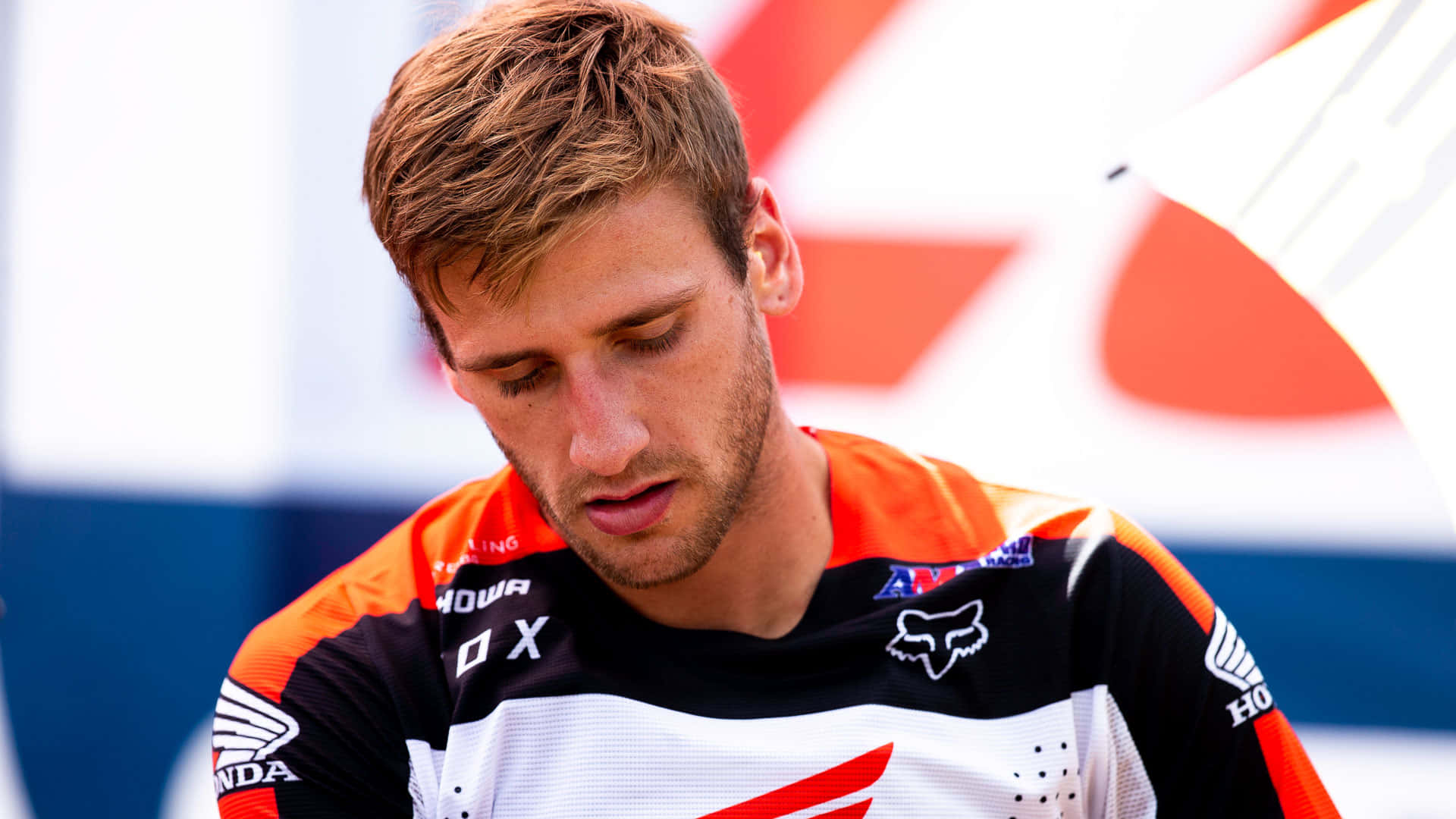 Ken Roczen 越野摩托賽車手桌面 壁紙 — 一位專注的越野電單車手，身穿橙色、黑色和白色裝備，在比賽中堅定地向下看。