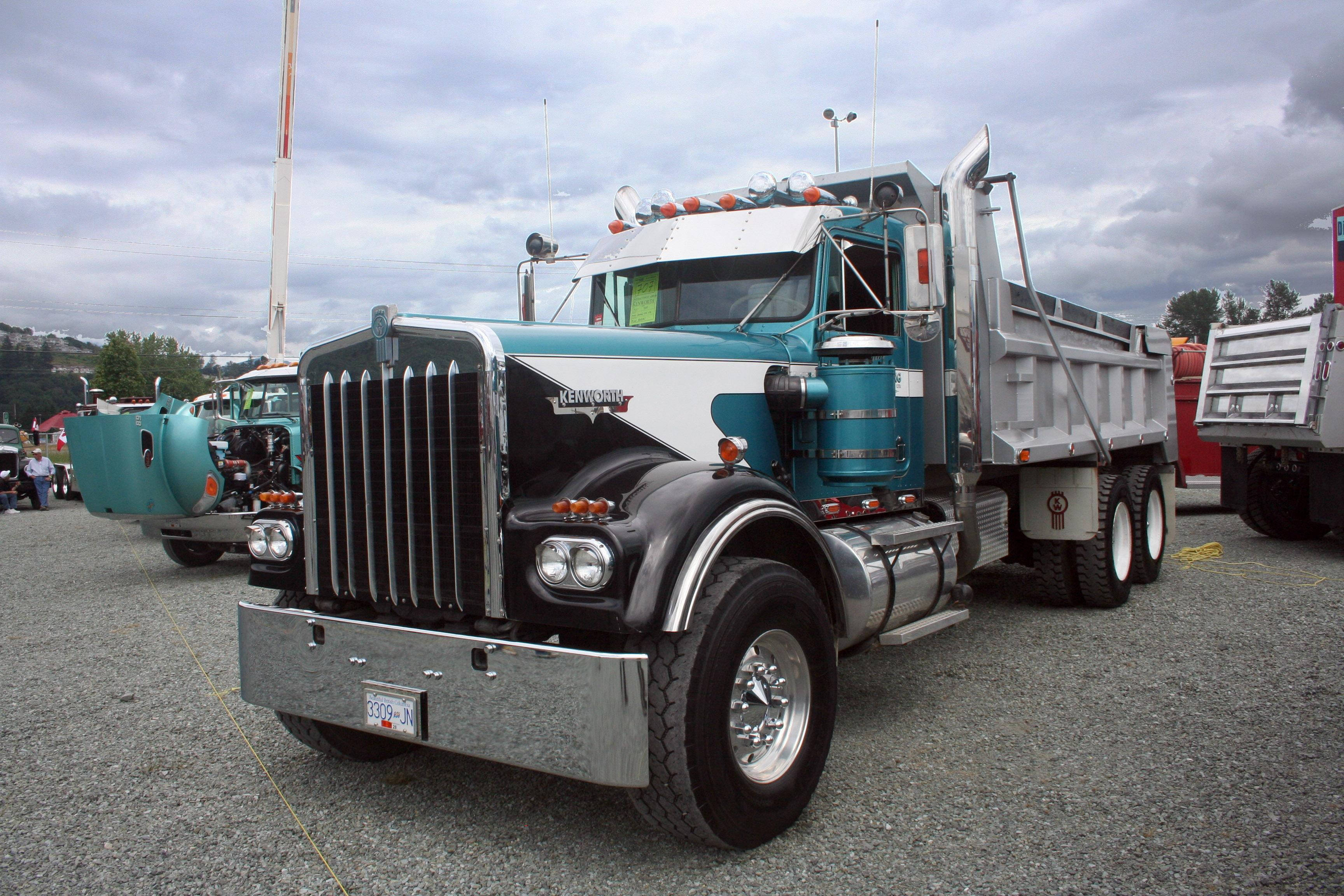 強勁Kenworth W900自卸卡車行駛中 壁紙 — 一輛帶有鍍鉻裝飾的藍綠色和白色Kenworth自卸車在陰天下的礫石上顯眼地停放。