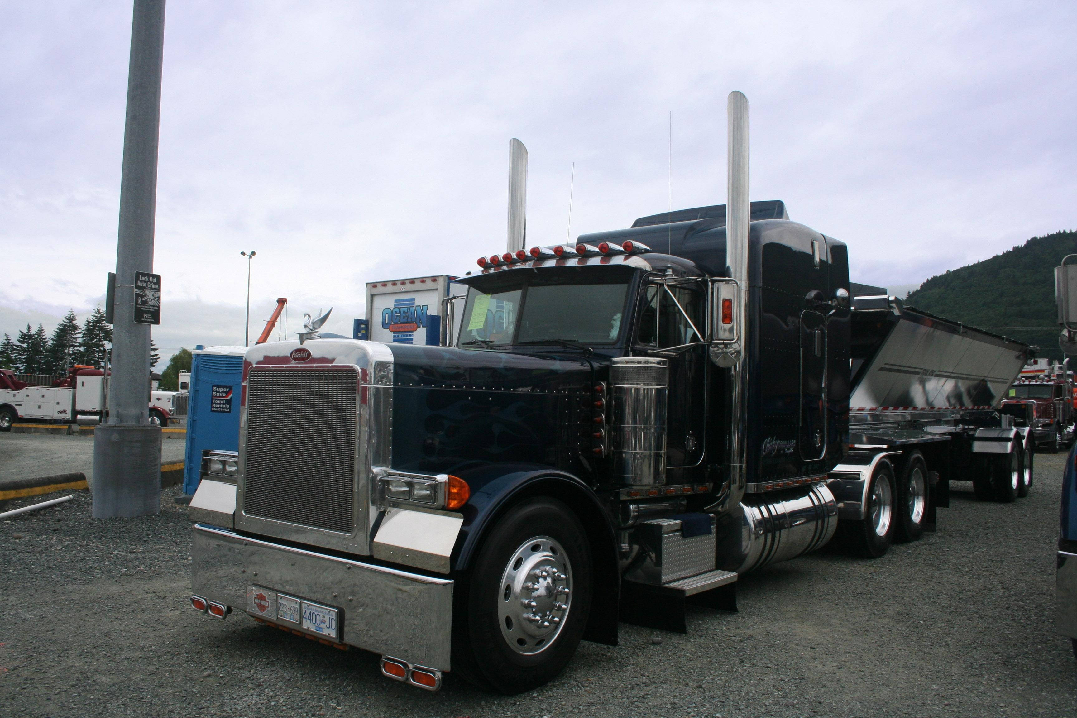 Kenworth W900鋼材運輸卡車 壁紙 — 一輛黑色Peterbilt半掛卡車，帶有鍍鉻裝飾和高排氣管，停在山附近的礫石上。